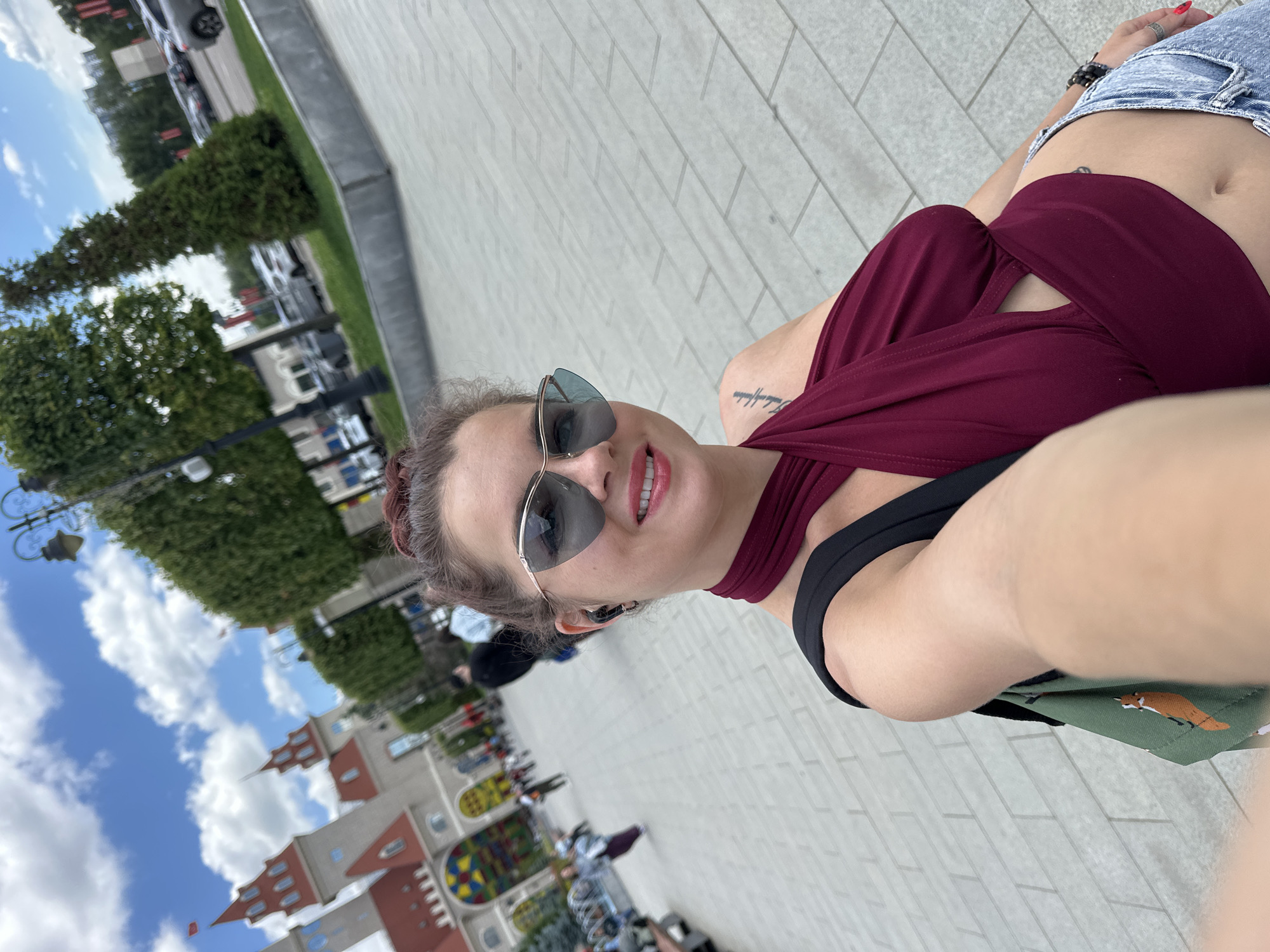 A woman is taking a selfie outdoors, possibly in Moscow, Russia. She is wearing a burgundy halter top, denim jeans, and cat-eye sunglasses. Her hair is pulled up in a bun, and she has a tattoo on her left shoulder. Behind her, there are some buildings that resemble castles, trees, streetlights, and parked cars. The ground is paved with light-colored tiles. The sky is partly cloudy. Several other people are visible in the background, walking and sitting.