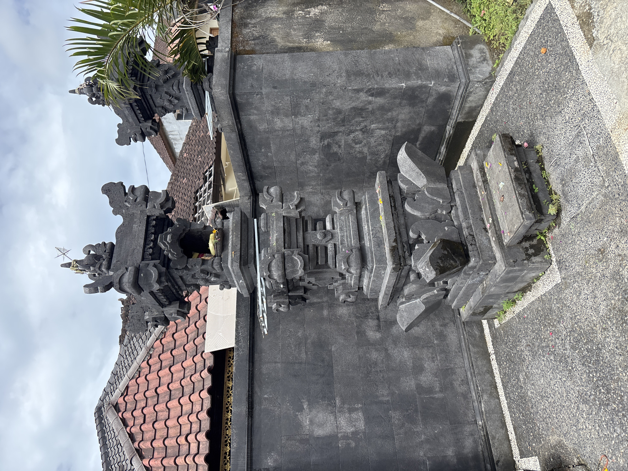 The image depicts a portion of a traditional Balinese architectural structure, likely a temple or a significant gate within a compound, in Kuta Selatan, Indonesia. The structure is predominantly composed of dark grey stone, exhibiting intricate carvings and layered ornamentation characteristic of Balinese design. Adjacent to the structure is a pathway or courtyard made of exposed aggregate concrete. In the background, a building with a tiled roof is partially visible, along with some vegetation. The lighting suggests an overcast day. No people are visible in the image.
