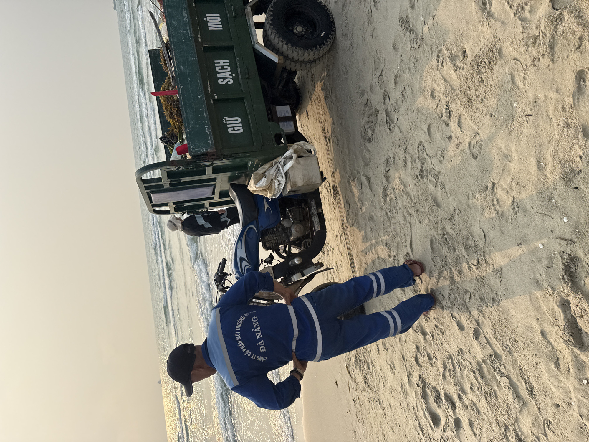 A man in a blue uniform with the words "Đà Nẵng" on the back stands next to a motorcycle parked on a sandy beach. He is wearing a black cap and sandals. A green truck with Vietnamese writing on the side is parked behind the motorcycle. The beach stretches out in the background with gentle waves visible. The location is likely a beach in Đà Nẵng, Vietnam, based on the writing on the uniform and the typical coastal landscape.