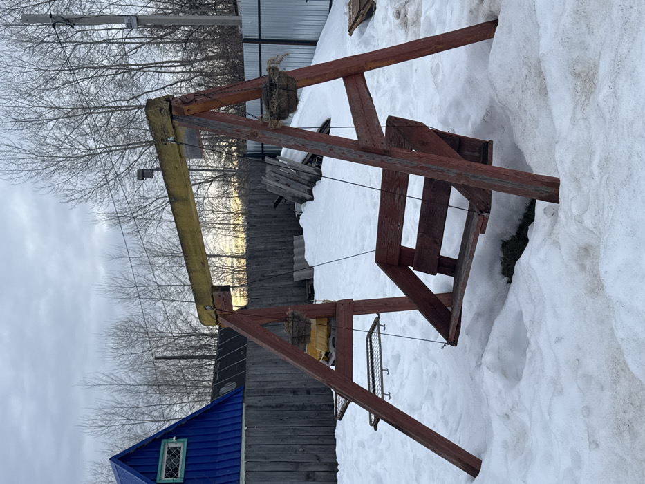 The image displays an outdoor scene in a residential or undeveloped area of Chaikovsky, Russia, during late winter or early spring. The foreground features a weathered wooden swing set with reddish-brown A-frame supports and an empty bench swing, and a yellow horizontal beam. Two metal grid shelves with hooks are attached to the left A-frame, while an empty, fibrous hanging basket is suspended from the right A-frame. The ground is covered in an uneven layer of melting snow, revealing patches of dirt and a small area of green grass beneath the swing. In the mid-ground, a dark wooden fence runs horizontally, separating the swing area from a cluttered space containing piles of wooden debris and corrugated sheets partially covered by snow. To the far left, a building with a blue pitched roof and a small, multi-paned window with green trim is partially visible. The background is dominated by numerous bare deciduous trees under an overcast, light grey sky. A utility pole with power lines is present on the far right, and a lighter-colored fence, possibly corrugated metal, is visible behind the swing set on the right side. No individuals are present in the scene.