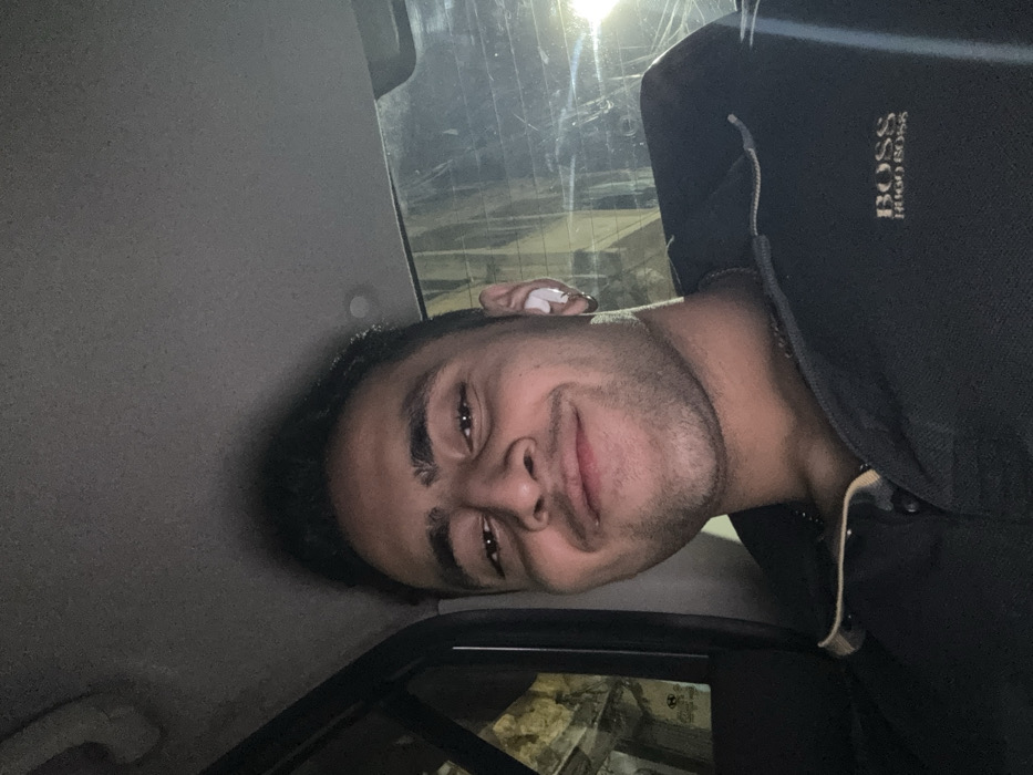 An image taken at night depicts a young adult male, appearing to be of South Asian descent, seated within a vehicle in Kolkata, India. The subject, positioned centrally and looking directly at the camera, exhibits dark hair, a mustache, and stubble. He wears a dark polo shirt with "BOSS Hugo Boss" embroidered on the left chest. A silver-colored chain is visible around his neck. A white earbud is present in his right ear, along with a silver hoop earring. The vehicle's interior headliner is gray. Through the blurred rear window, streetlights and indistinct building structures characteristic of an urban environment are faintly visible. A partial "Hyundai" logo is discernible on the left side of the rear window.