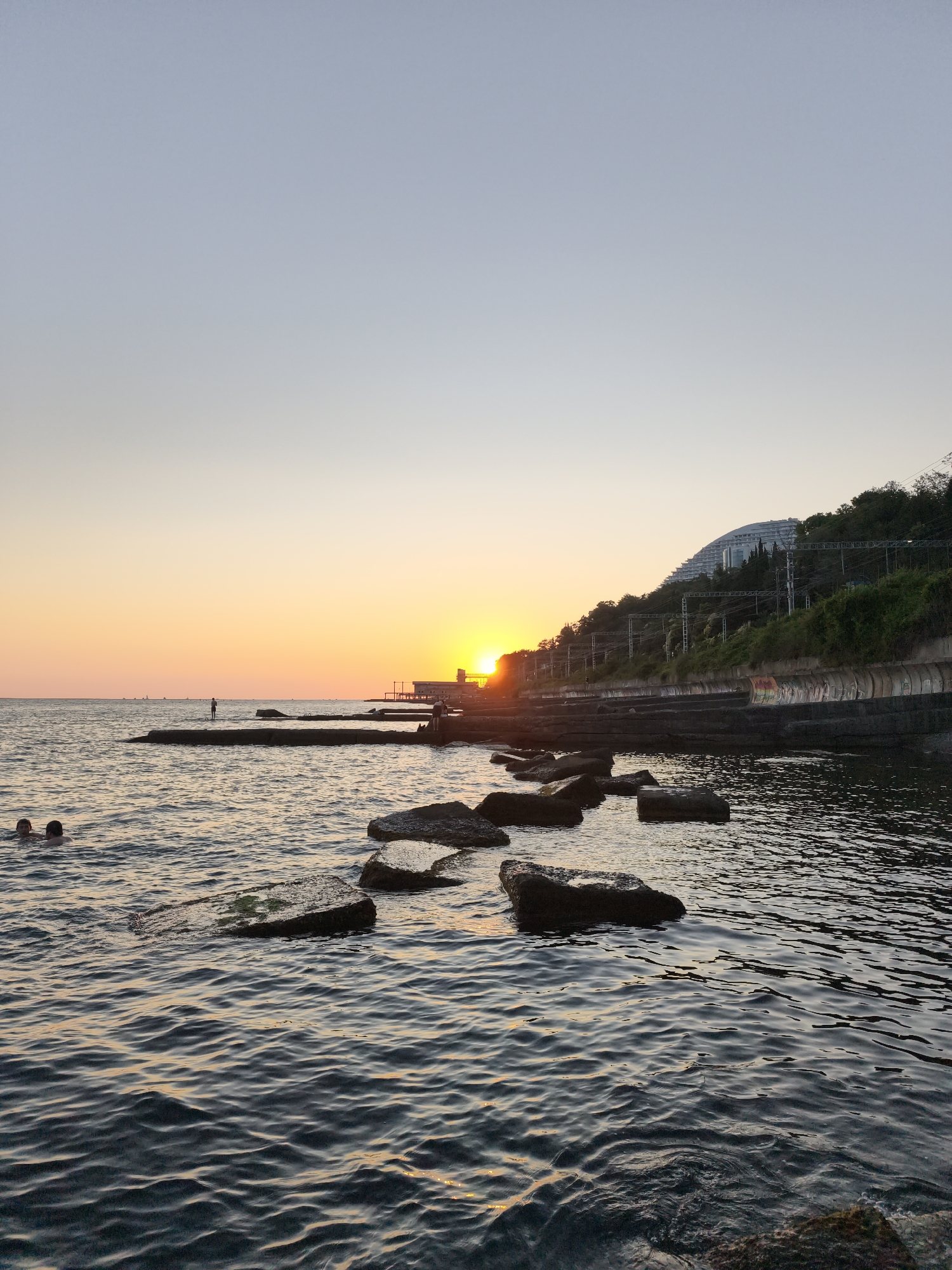 This is a vertical, eye-level, medium shot taken during sunset at a coastal area in Sochi, Russia. The sun is low on the horizon, casting a warm orange glow across the sky and reflecting on the water. In the foreground, the dark, rippling water of the Black Sea fills the lower half of the frame, with several dark, flat rocks partially submerged. Two people are visible swimming in the water on the left side of the image. In the middle ground, a concrete pier extends into the water, with a single person standing on it. Further back, another section of a concrete pier is visible, along with a building and a hill covered in green trees and vegetation. A large, modern building with a curved roof is situated on top of the hill, and a railway track runs along the hillside. The overall scene depicts a serene evening at the beach.