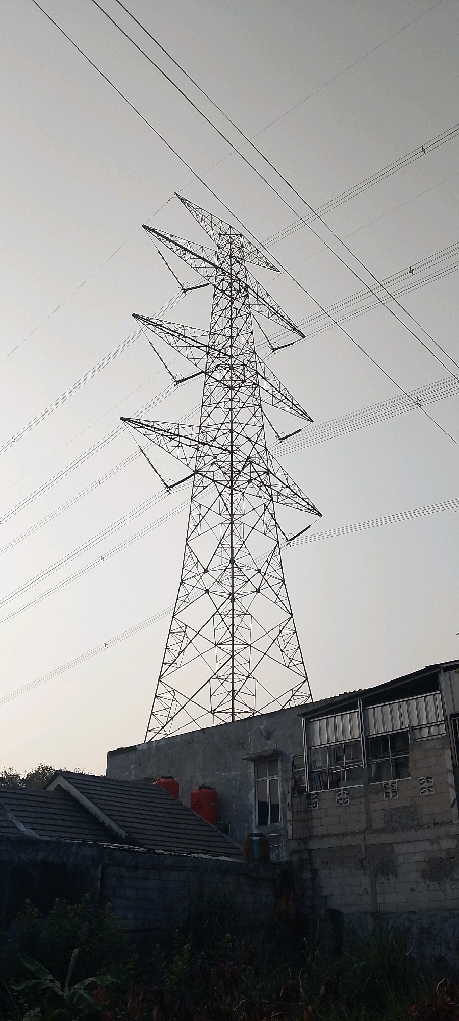 The image shows a large, lattice-style electricity pylon dominating the center of the frame, with multiple power lines stretching across the sky. The pylon is silhouetted against a pale, overcast sky. At the base of the pylon, on a raised concrete platform, are several buildings. To the left is a house with a dark grey tiled roof and two bright orange water tanks visible on top of it. To the right is a more industrial-looking building constructed of grey brick and metal paneling, with multiple windows. The foreground is filled with dark green foliage and some dry grass, obscuring the ground level. The scene appears to be located in a semi-urban or industrial area in Satriamekar, Indonesia, likely on a cloudy day. There are no people visible in the image.