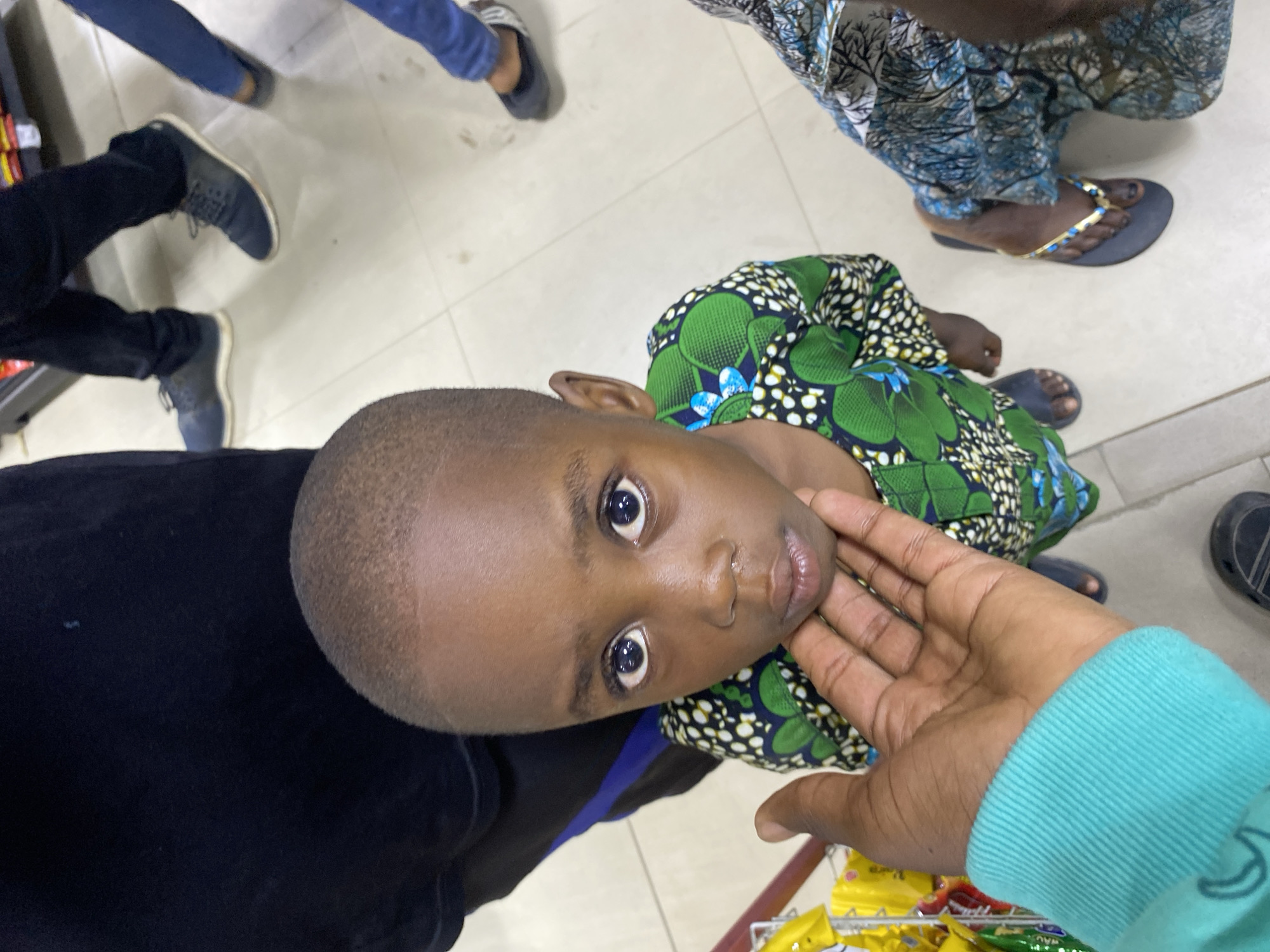 A small child is looking up at the camera. The child is wearing a green and black patterned shirt. A hand is touching the child's chin. The child's eyes are wide and expressive. The background shows other people's legs and feet. The environment appears to be a store. The image was likely taken in Lagos, Nigeria.