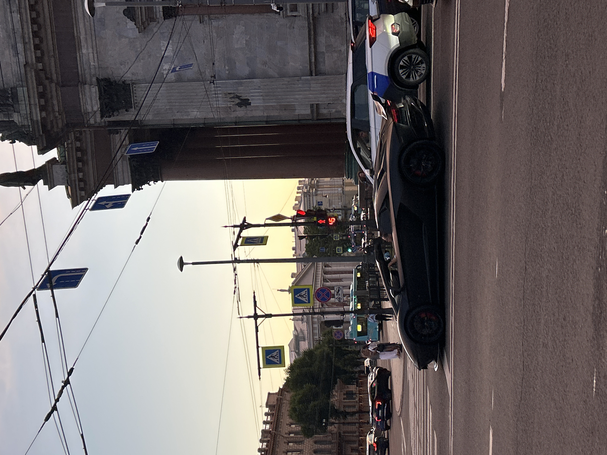 The image depicts a street scene in a city, likely during the daytime with soft lighting suggesting late afternoon or early morning. In the foreground, a black convertible sports car is stopped at a pedestrian crossing. Behind it, a white SUV is also stopped, and another vehicle is partially visible to its right. Further in the background, a light blue bus is seen, along with several other cars and pedestrians. Various traffic signs are visible, including pedestrian crossing signs and directional signs. The architecture in the background suggests a European city, with large, ornate buildings and overhead electrical wires for what appears to be a trolleybus system. The sky is clear with a pale gradient, indicating a tranquil atmosphere.