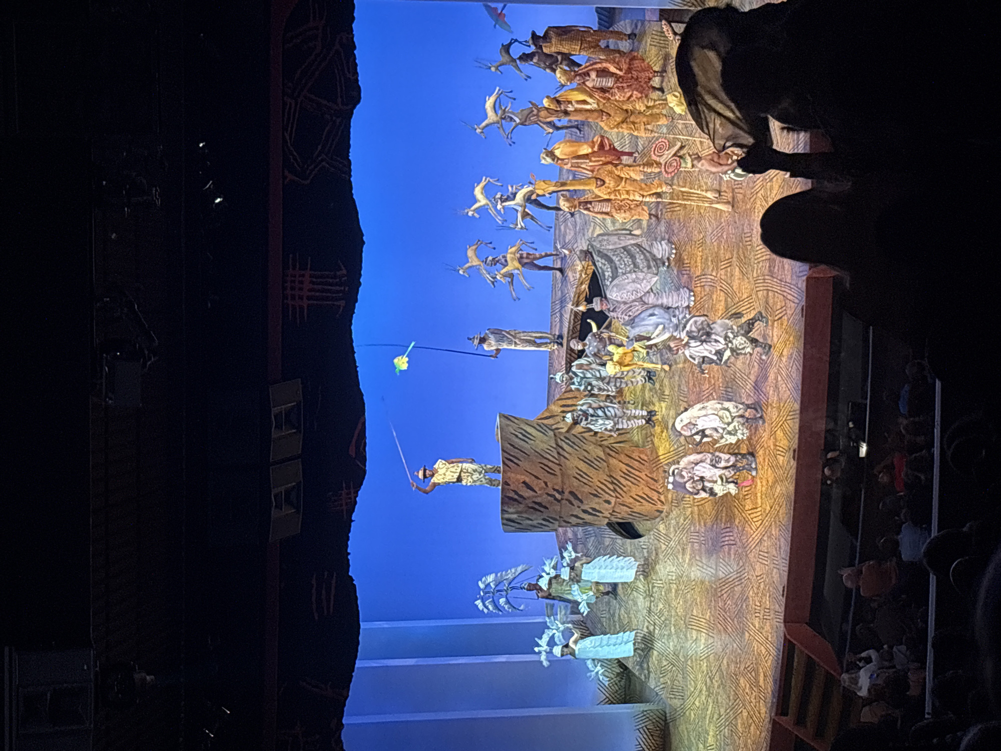 The image depicts a scene from the musical "The Lion King," likely performed at the Minskoff Theatre in New York City. The stage is set with a vibrant blue backdrop and intricately designed costumes and puppets representing various animals of the African savanna. A performer stands atop a prop, holding a fishing rod with a dangling element, while others are positioned around the stage, representing various animals, including gazelles and zebras. The audience is visible in the foreground, with heads and shoulders silhouetted against the dimly lit theater.