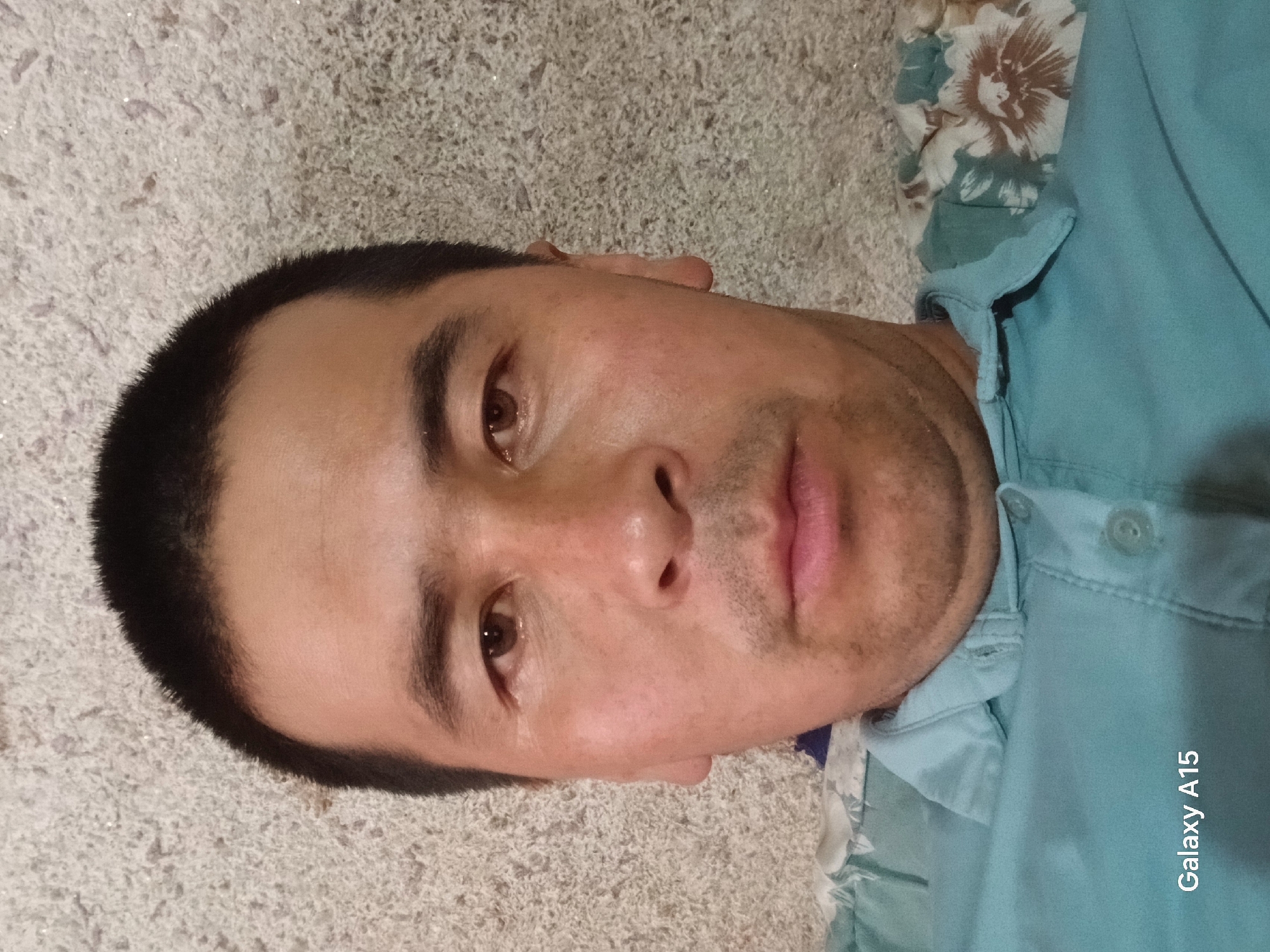 A selfie of a man is taken from the chest up. He has short, dark hair and brown eyes. He is wearing a light blue collared shirt. The background is a textured, light-colored surface. The Samsung Galaxy A15 logo is visible in the bottom left corner.