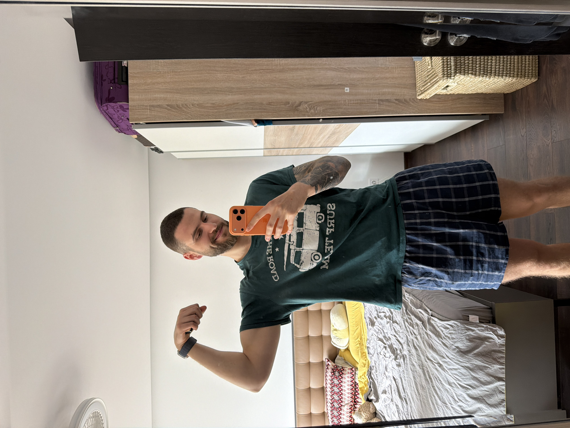 A man is taking a selfie in a mirror. The man is wearing a dark green t-shirt with a white graphic and plaid shorts. He is flexing his left arm and holding an orange smartphone in his right hand. The room contains a bed with a gray comforter, a tan headboard, and decorative pillows. To the left of the man is a white wall. A wardrobe with a black top, a light brown middle section, and a white bottom is visible in the background. A wicker basket and shoes are on top of the wardrobe. The floor is dark wood. The city of Sofia, Bulgaria, is not discernible in the image.