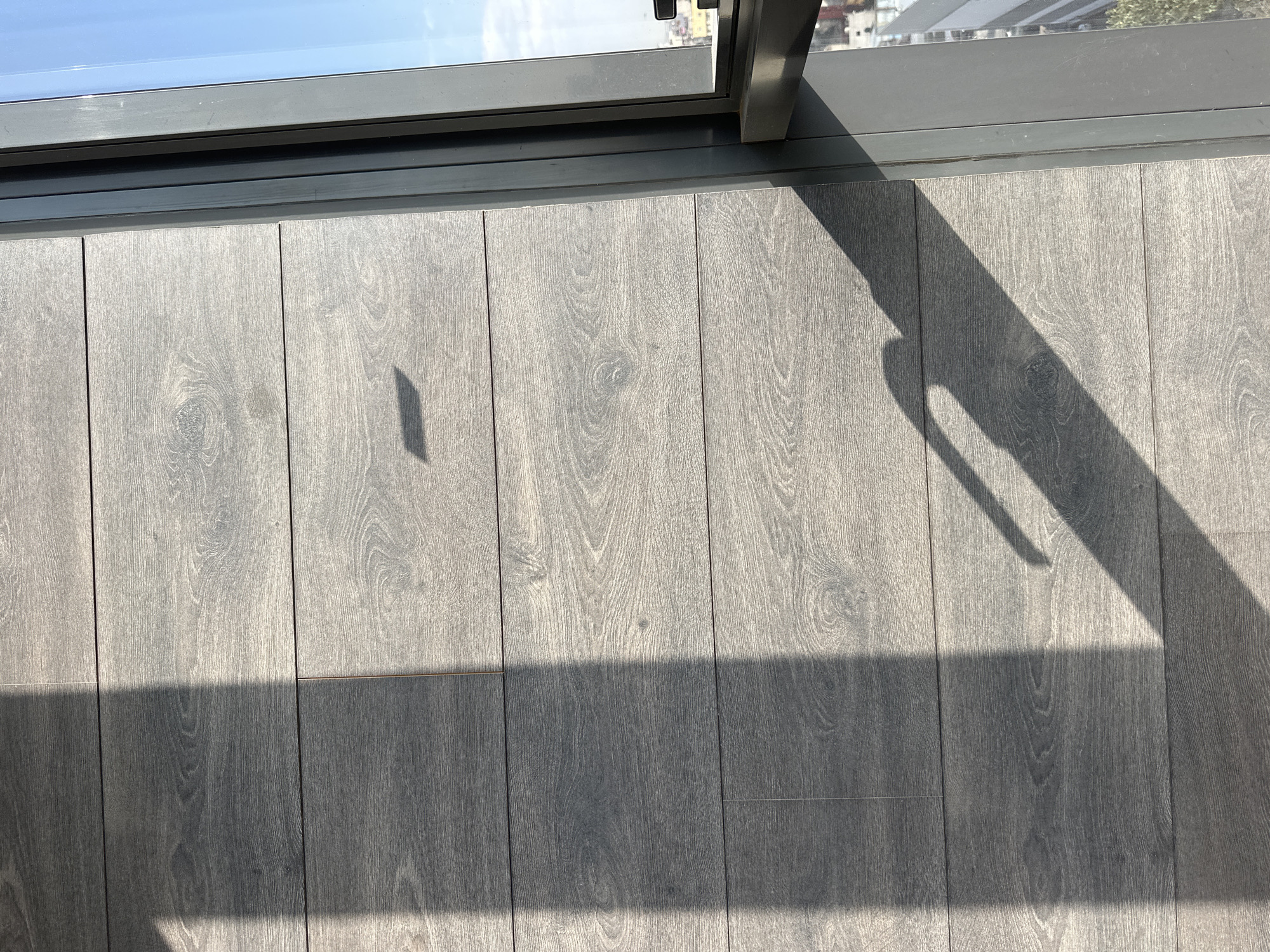 The image depicts a section of gray wood flooring next to a window with a metal frame. The window offers a partial view of a building and the sky. A shadow cast by the window frame and handle falls across the flooring. The floorboards are aligned vertically in the image. The location is inferred to be Huixquilucan de Degollado, Mexico.