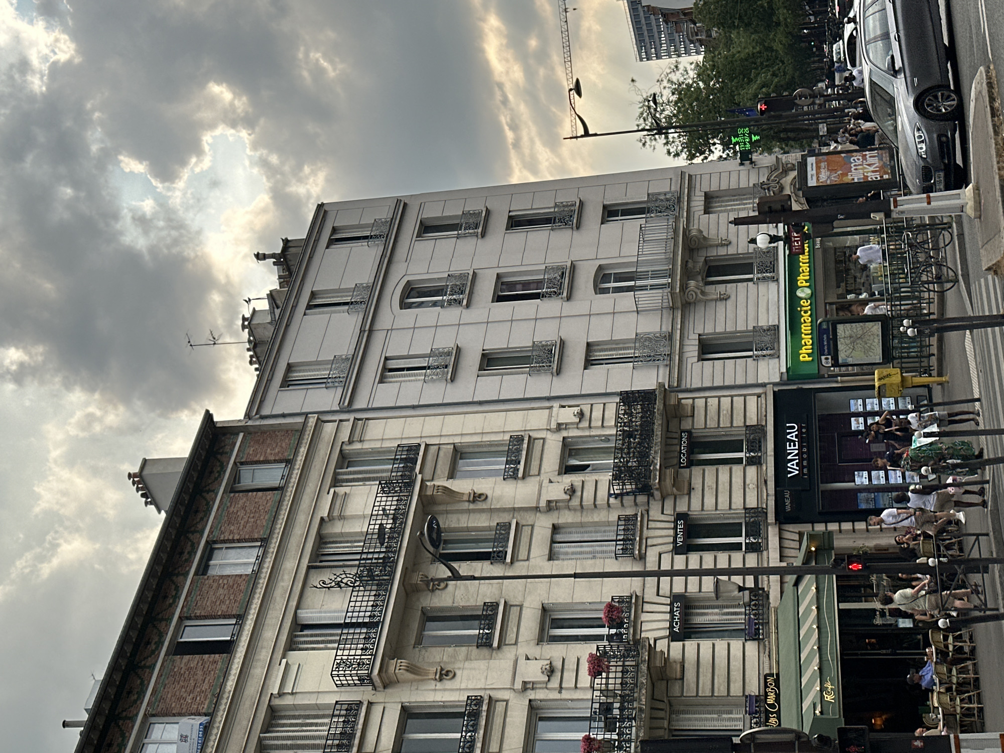 This image captures a street scene in Paris, France, under a cloudy sky. The foreground features a light-colored car driving on the right side of the road. To the left, a building displays storefronts including "R café" with an awning and outdoor seating, and "VANEAU immobilier" with posters displayed in the windows. Further to the left, a green awning covers a pharmacy labeled "Pharmacie Pharm". The buildings are multi-story structures with ornate balconies and windows. Several people are visible walking on the sidewalks and sitting at outdoor cafe tables. A traffic light displays a red signal. A lamppost stands prominently in the middle of the scene. In the background, trees and other buildings can be seen, including a crane in the distance, suggesting ongoing construction. The overall impression is of a bustling urban environment.