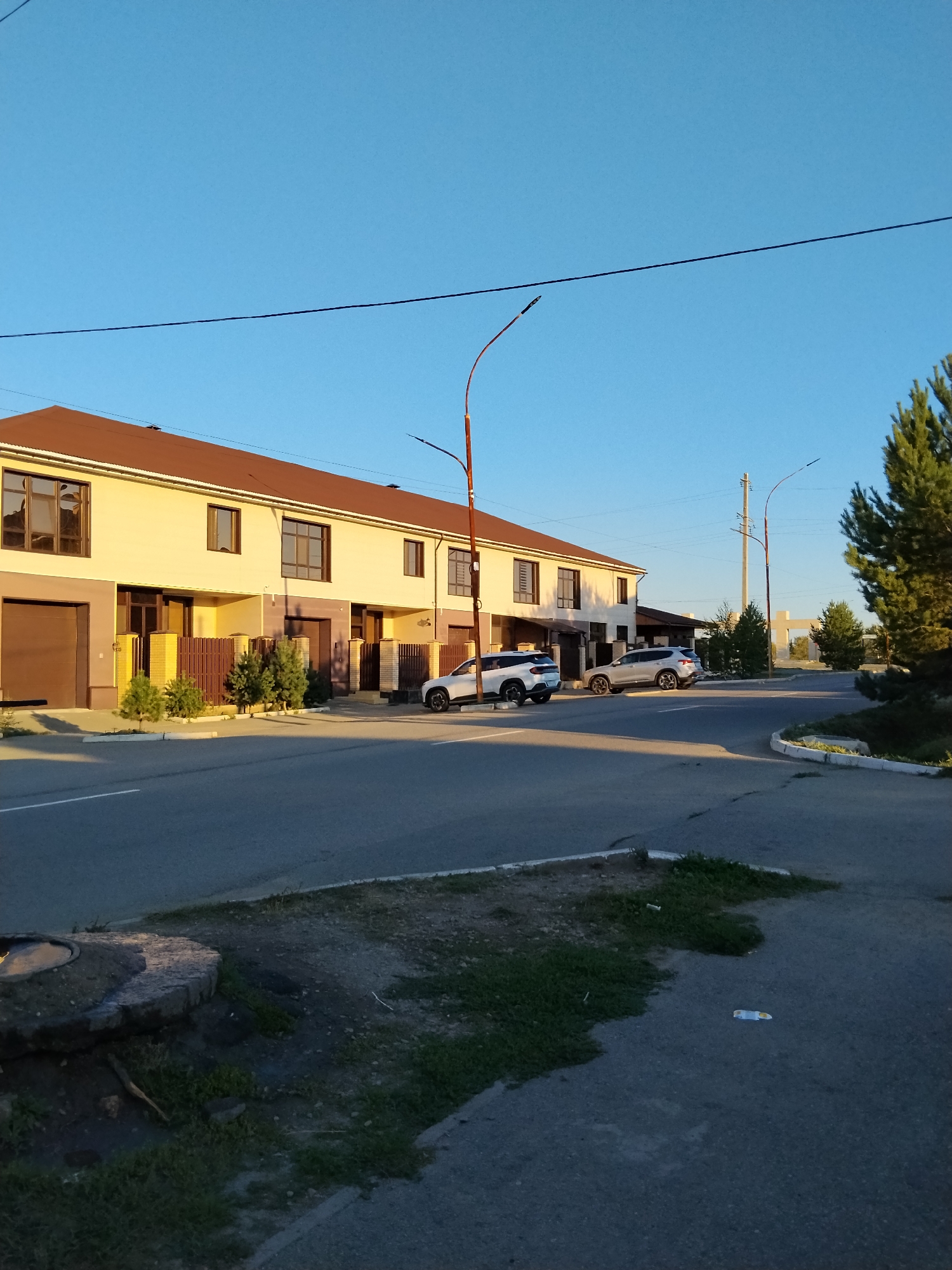 A row of modern, beige townhouses with brown roofs and garages lines a paved road in Kokshetau, Kazakhstan. Two white SUVs are parked on the street in front of the townhouses. A brown wooden fence separates the properties from the road. Small, manicured evergreen trees are planted in front of the fence. The sky is clear and blue, and the sun is casting long shadows, suggesting it is late afternoon or early evening. Power lines are visible strung between utility poles. On the right side of the image, a tall evergreen tree stands next to the road. In the foreground, a concrete structure, possibly a manhole cover or wellhead, is visible on the grassy verge. The overall scene depicts a residential street in a suburban area of Kokshetau.