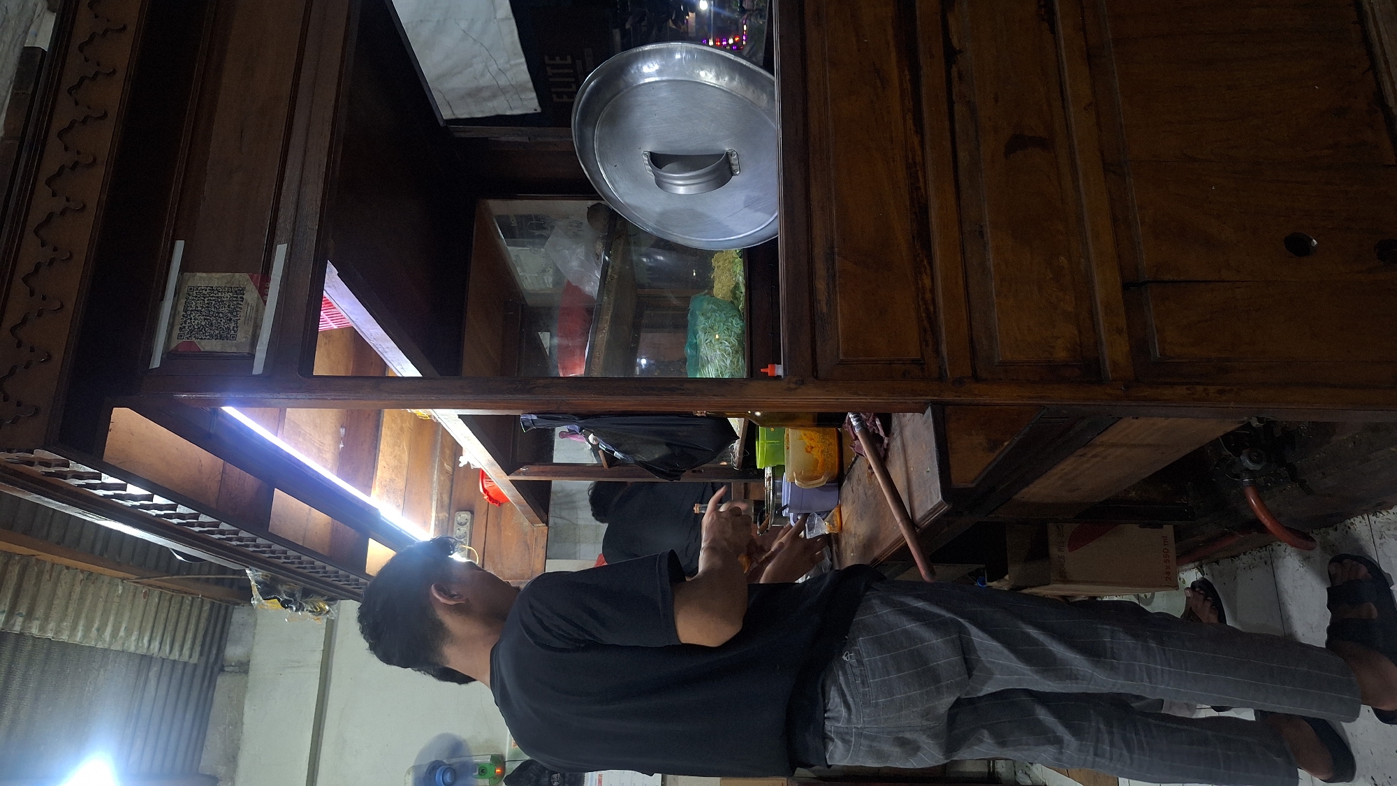 A man in a black t-shirt and grey plaid pants stands facing a wooden cabinet, presumably preparing food. The cabinet has glass-enclosed shelves containing food and supplies, including a metal lid and various containers. The location is likely a food stall or small eatery in Sukaharja, Indonesia, given the typical street food setup.
