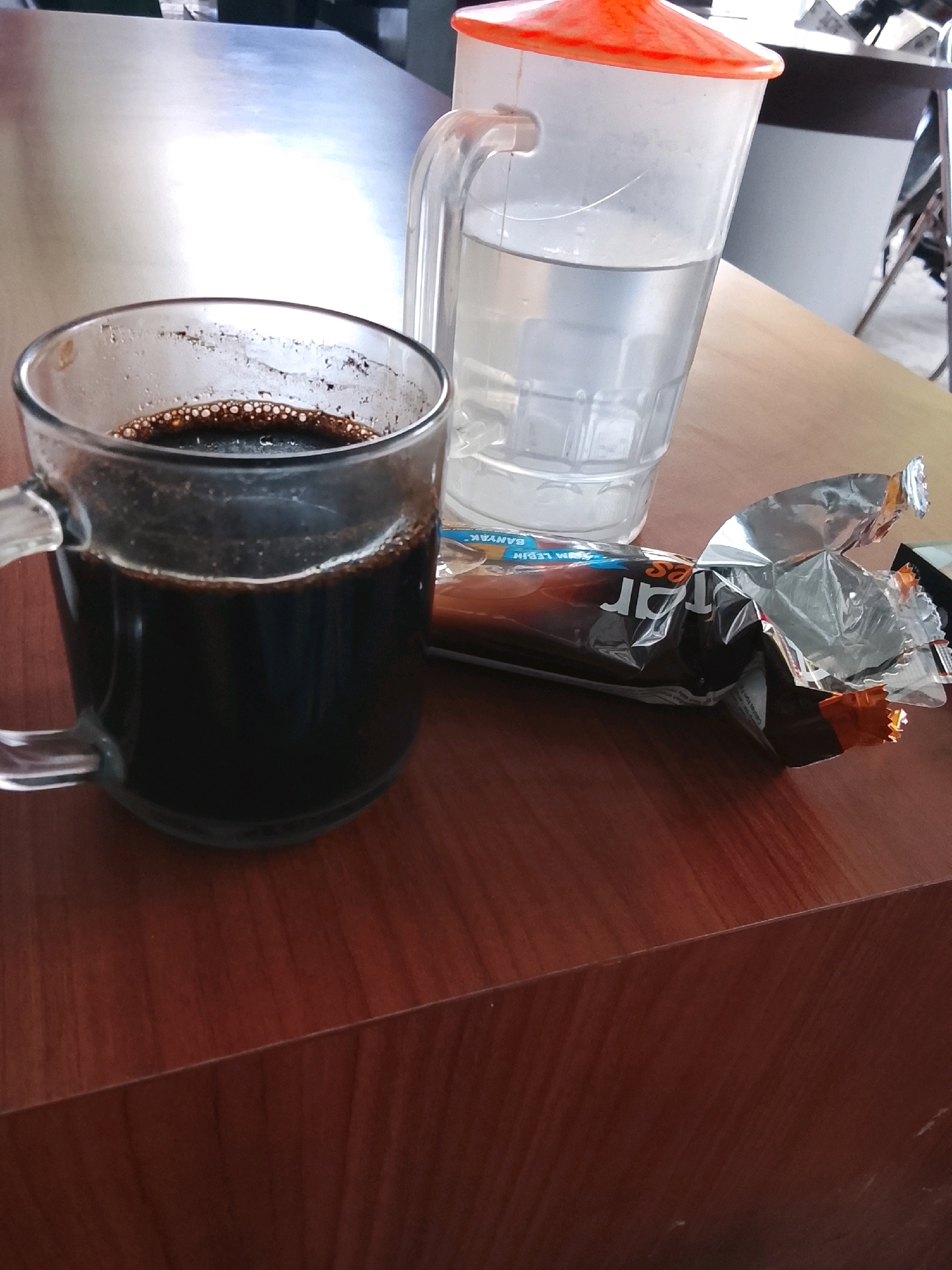 This image depicts a close-up, eye-level view of a wooden table at a cafe in Slawi, Indonesia. The scene is set indoors, likely in the morning, given the lighting.
Dominating the foreground is a clear glass mug filled with dark coffee. The coffee has a frothy layer on top, suggesting it was recently poured or brewed. The mug has a prominent handle on its left side.
To the right of the coffee mug, a partially unwrapped candy bar or snack is visible. The wrapper is silver and orange, with some text in a foreign language, possibly Indonesian, hinting at its origin or brand. The wrapper is crumpled, indicating the product has been opened and possibly partially consumed.
In the background, to the right of the coffee mug and behind the snack, stands a clear plastic pitcher filled with water. The pitcher has a bright orange lid. The pitcher is positioned on the same wooden table.
The table itself is made of dark wood with a visible grain pattern, suggesting it is a sturdy, perhaps varnished surface. The overall ambiance of the image is casual and relaxed, suggestive of a moment of respite. The background is slightly blurred, but hints of other tables and chairs suggest the presence of a larger dining area or cafe setting. The lighting appears to be natural, coming from an unseen window or overhead light source, casting soft shadows on the table.