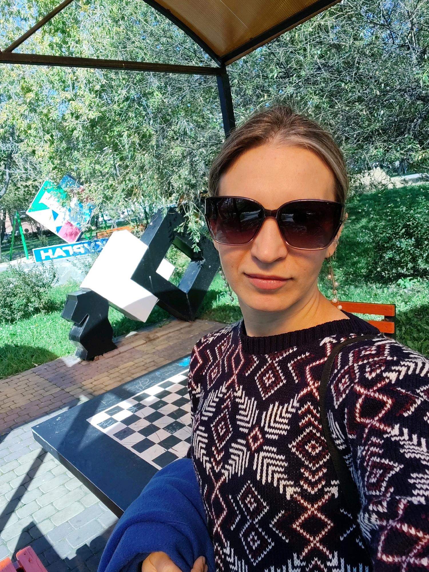 A woman, facing the camera with sunglasses, poses for a selfie outdoors. She is wearing a dark blue sweater with a geometric pattern in white and burgundy. She has her hair pulled back and is wearing long earrings with pearls. Behind her, a large black and white sculpture resembling a geometric puzzle is visible, along with a checkerboard pattern embedded in a bench or table. Trees with green foliage and a blue sky are in the background. The scene appears to be in a park or public outdoor space.