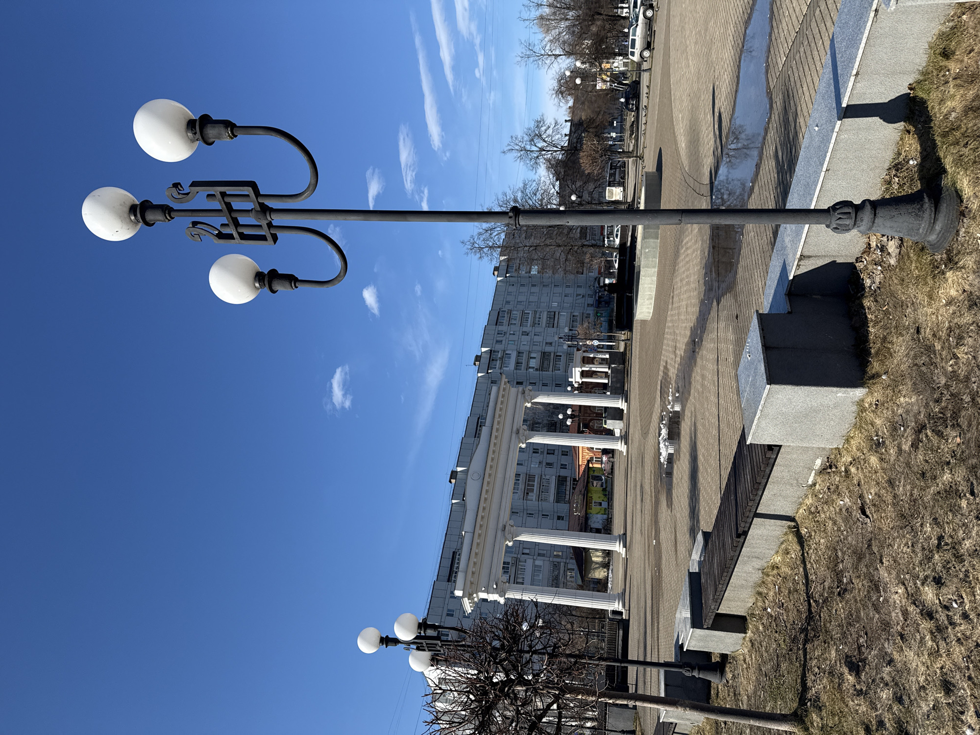 The image shows a street scene in an unidentified city. In the foreground, there are lampposts with three spherical white lights each. The lampposts are made of dark metal with ornate designs. The sky is blue with some wispy clouds. In the background, there is a building with white columns, partially obscuring a gray apartment building. The street is paved with brick and lined with a low concrete wall. There is grass and some bare trees on the right side of the image. A few vehicles are parked in the distance.
