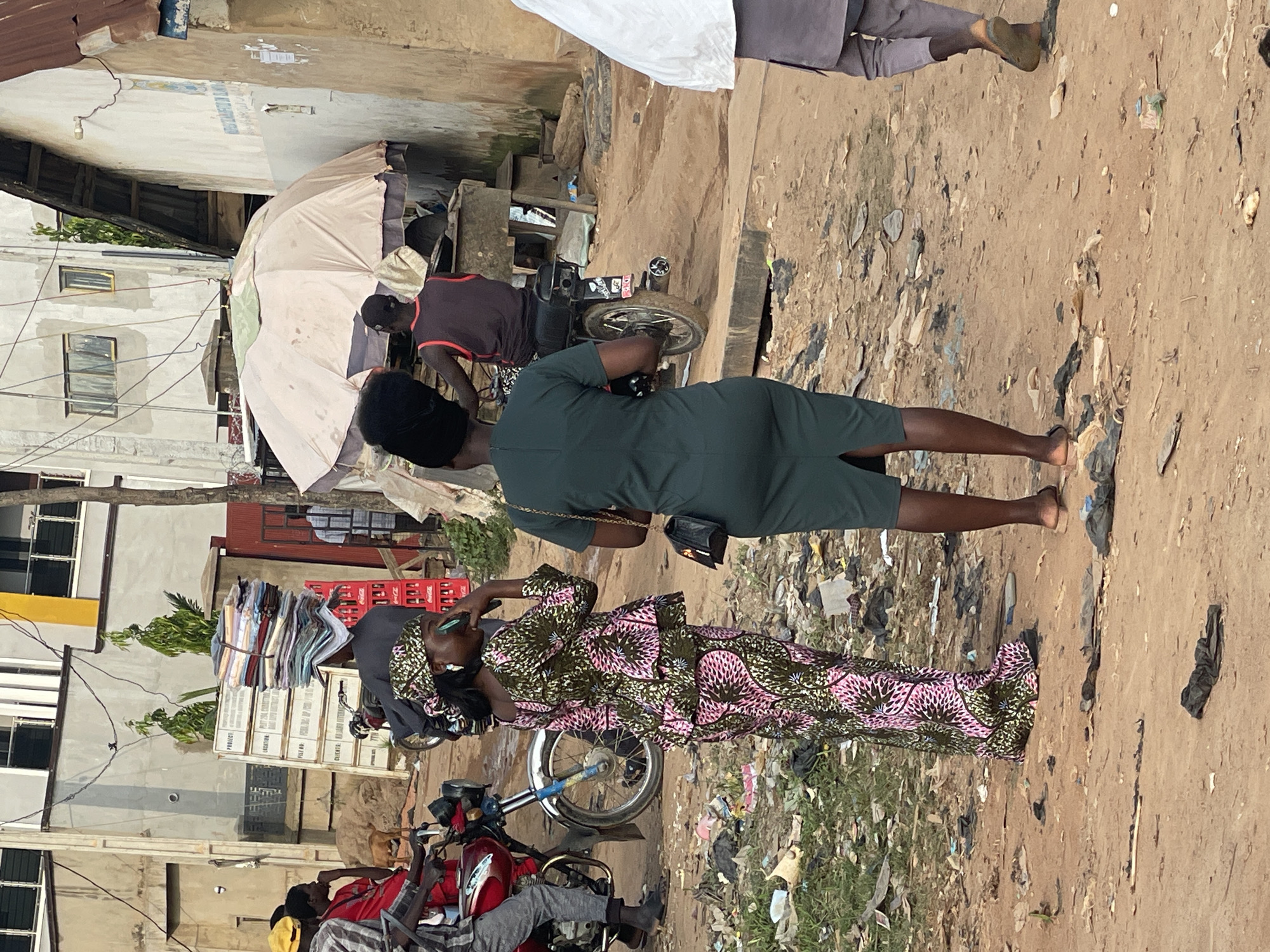 The image depicts a bustling street scene in Abuja, Nigeria, characterized by its dusty, unpaved ground littered with debris. In the foreground, two women are prominent. The woman on the left, adorned in a vibrant pink and green patterned traditional outfit and headscarf, is captured mid-conversation, holding her phone to her ear. To her right, a woman in a dark green dress stands with her back to the camera, carrying a black purse.
Behind them, a motorbike rider wearing a dark sleeveless shirt is visible, adding to the dynamic atmosphere of the street. Further in the background, stacks of folded fabrics are being transported, balanced precariously on a person's head, highlighting the commercial activity of the area. A large, weathered umbrella provides shade over a stall or vendor setup, suggesting a marketplace or informal trading post. The surrounding buildings, with their neutral-toned facades, contribute to the urban backdrop of this Nigerian city. The overall impression is one of a lively, everyday street in Abuja, where commerce, transportation, and daily life intersect.