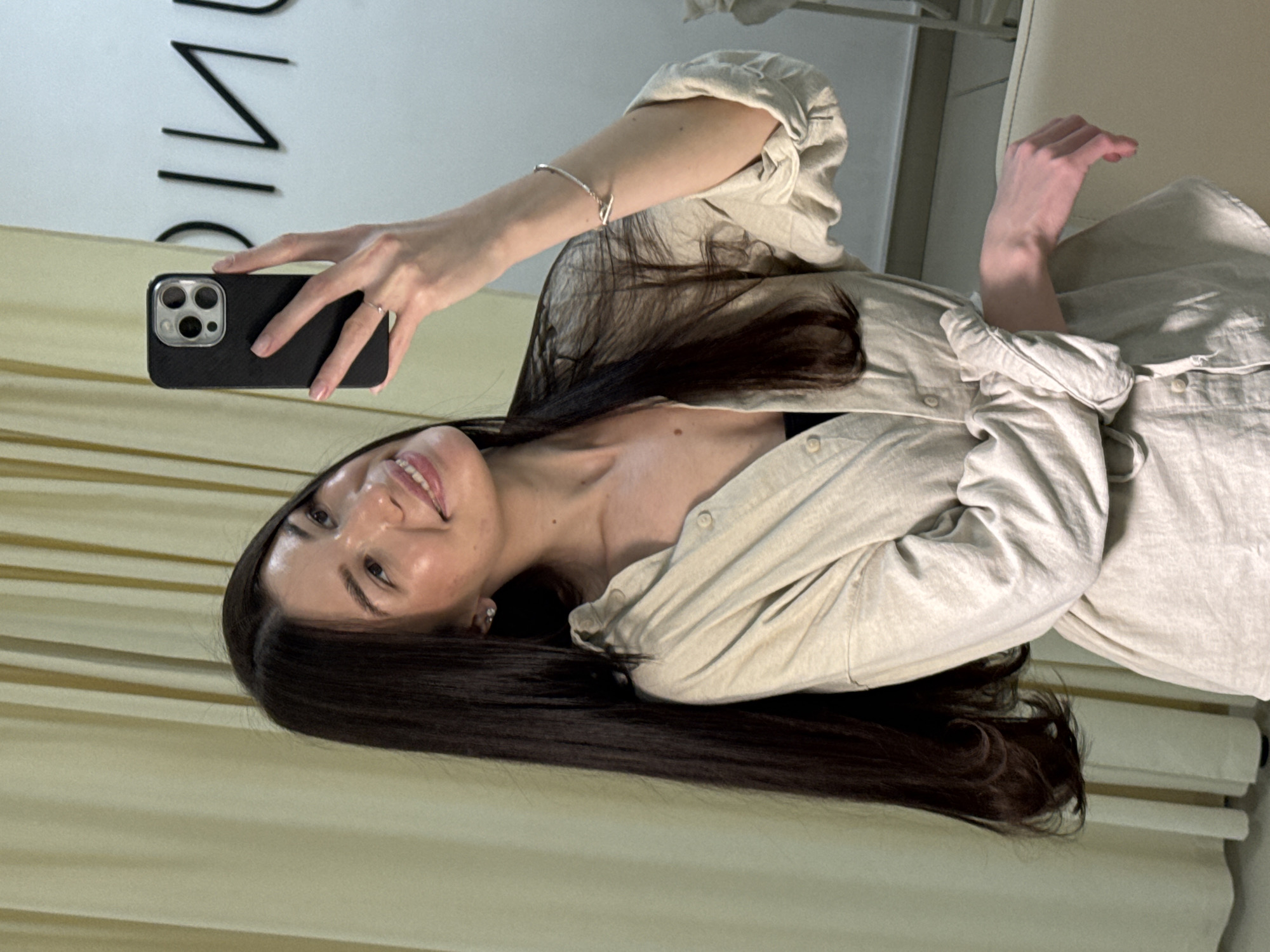 A woman with long, dark hair is taking a selfie with a black iPhone in what appears to be an indoor setting. She is wearing a light-colored, button-down shirt. There is a sign or lettering visible in the background. The setting appears to be in a room in the city of Chaikovsky, Russia.