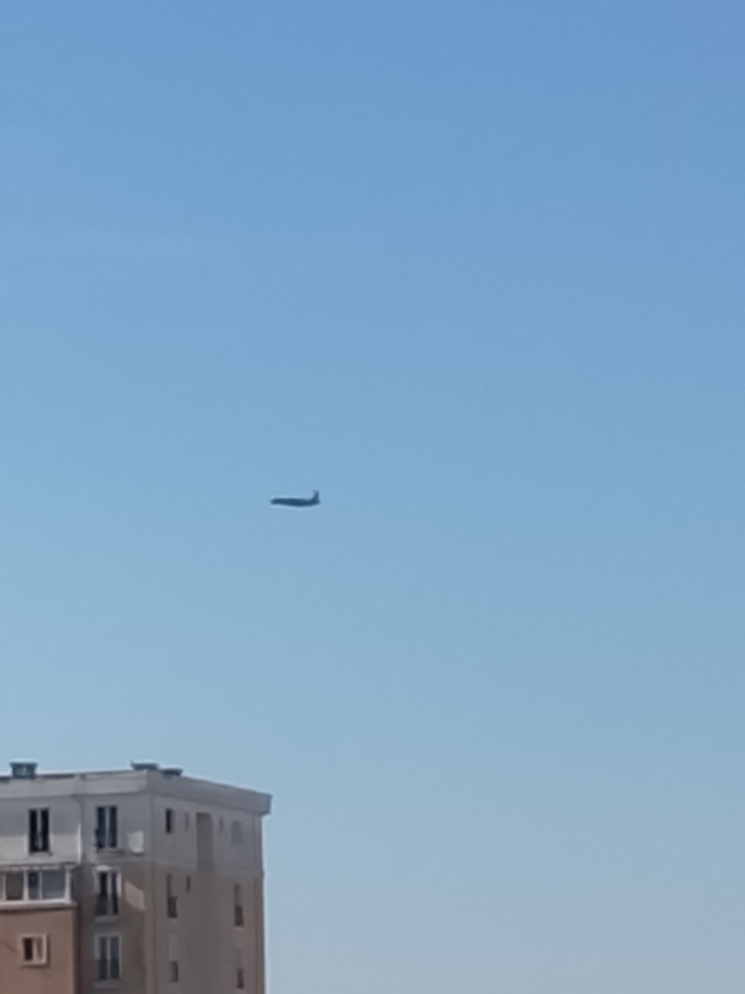 An airplane is visible in the clear blue sky above a building in Crețuleasca, Romania. The airplane is a dark silhouette against the sky. The building is light-colored with visible windows and appears to be an apartment complex. The image is taken from a low angle, looking up at the sky.