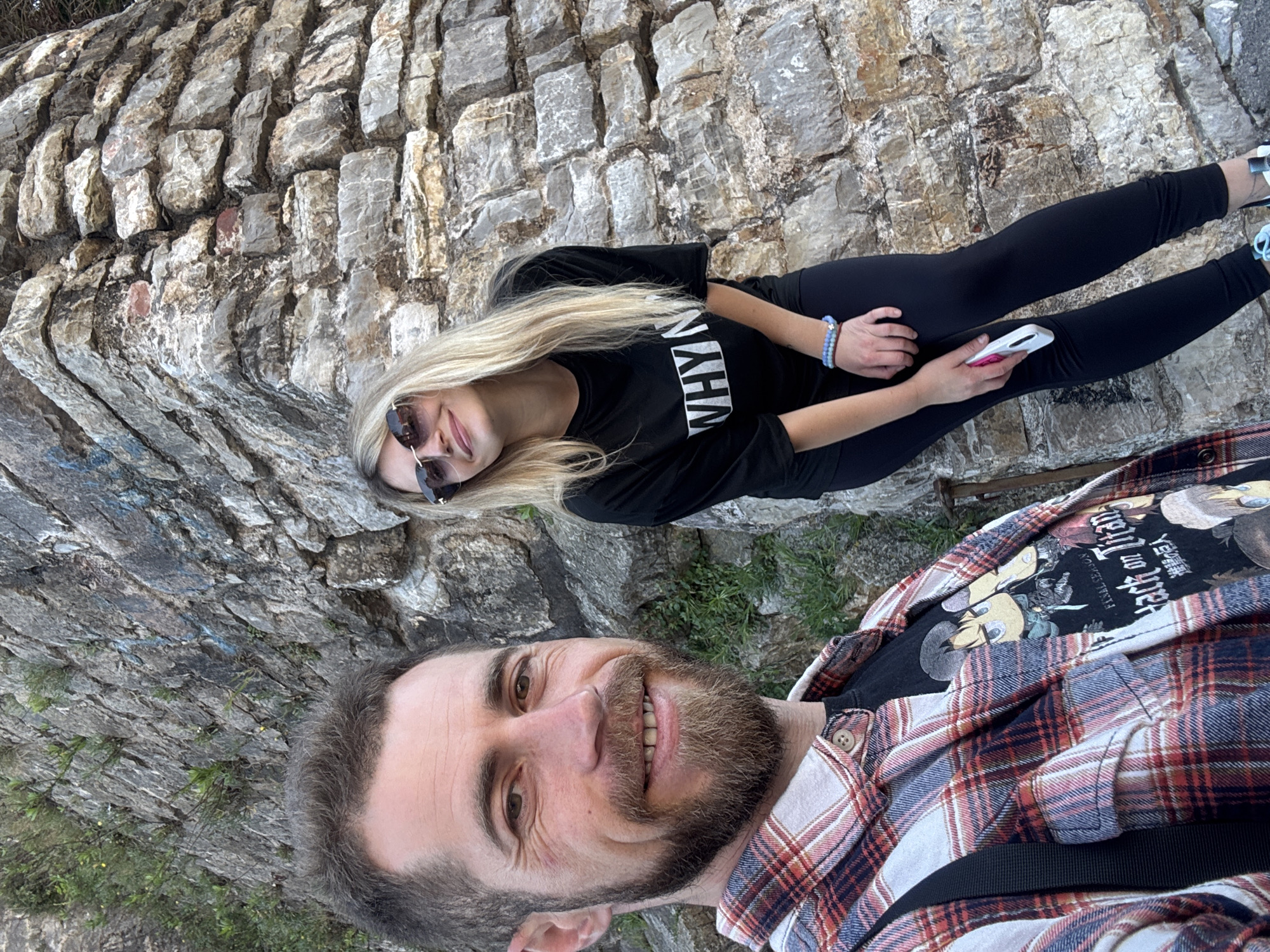 A medium shot depicts a Caucasian male and a Caucasian female posing for a selfie in Budva, Montenegro. The male, located in the foreground, is smiling at the camera; he has short brown hair, a beard, and is wearing a red and white plaid shirt over a black t-shirt. The female is positioned behind the male, lying on her side. She is blonde, wearing sunglasses and a black t-shirt with the word "WHYN" printed on it. She is holding a white smartphone. Both are positioned in front of a gray stone wall. The photo is taken at an angle.