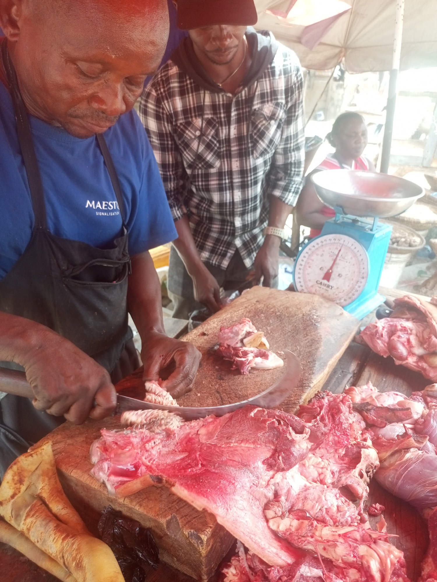 At a butcher's stand in Sabon Tasha, Nigeria, a man in a blue shirt and black apron is cutting meat on a wooden block with a large knife. Another man in a plaid shirt stands behind him, and a woman sits behind them with a blue scale. Raw meat is piled on the cutting surface.