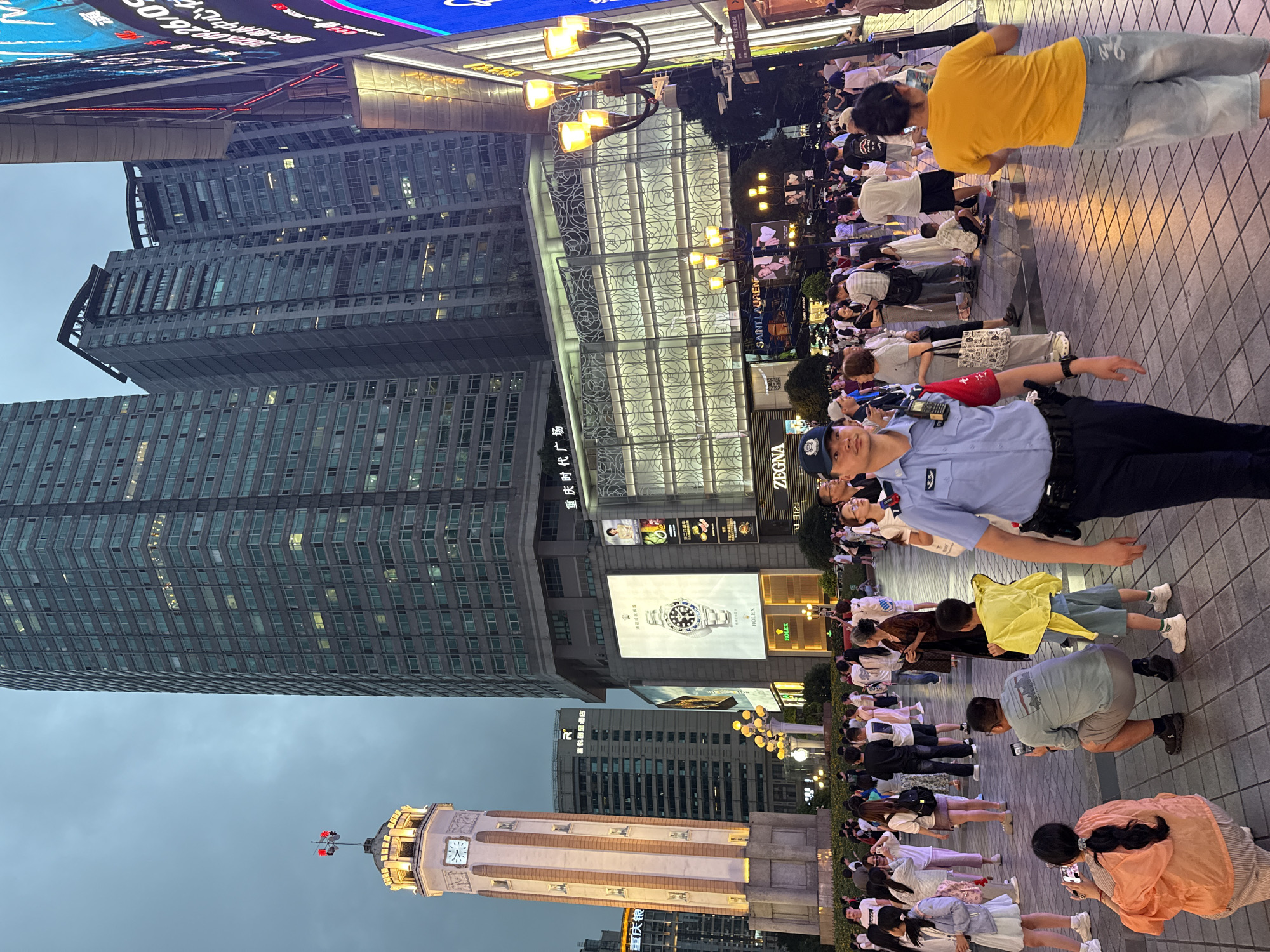 The image depicts a bustling street scene in Yuzhong District, China, with a prominent clock tower and modern skyscrapers forming the backdrop. The sky is overcast, suggesting it is evening or a cloudy day. A diverse crowd of people, including families, individuals, and a police officer, are present in the foreground and middle ground, navigating the paved walkway.
The clock tower, a central feature, is illuminated and displays a clock face. Various shops and advertisements are visible on the lower levels of the surrounding buildings. One large advertisement features a watch, identified as a Rolex. Another displays the brand Zegna. Illuminated street lamps line the walkway, adding to the evening ambiance.
People are engaged in a variety of activities. Some are walking, while others are standing or sitting. A man in the center is crouching, taking a photograph with his phone, and a young boy stands beside him. A woman in an orange top is also crouched down, seemingly taking a photo. A police officer in uniform is walking towards the viewer on the right side of the frame. The clothing styles of the people suggest a casual atmosphere. The overall impression is that of a lively urban public space.