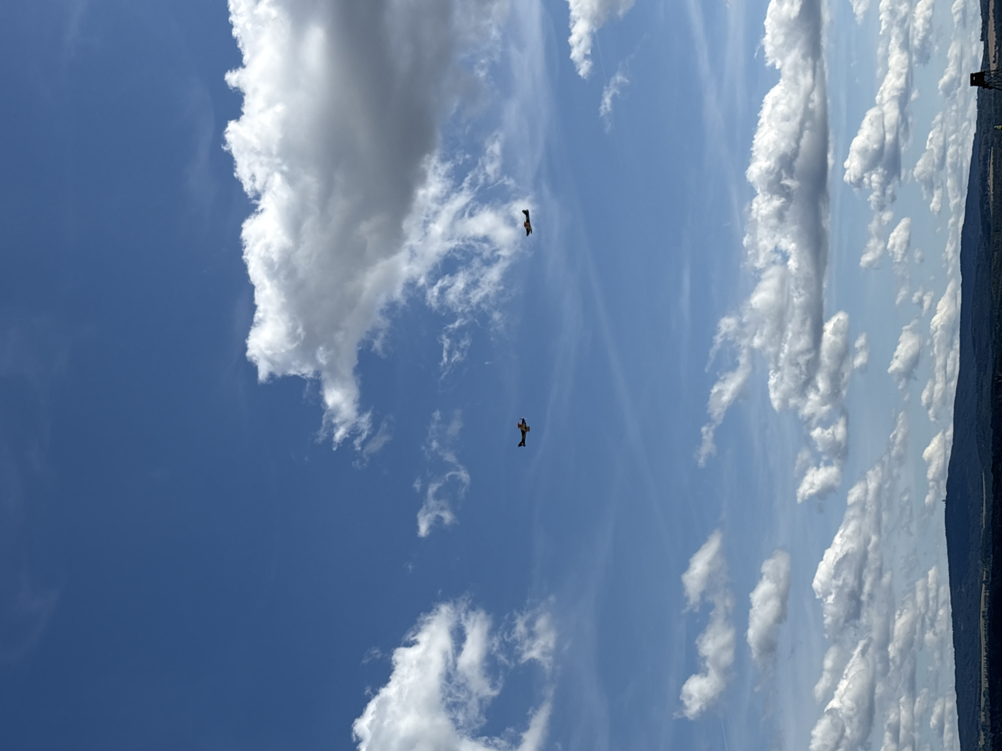 This image captures a wide-angle view of a bright blue sky filled with scattered white and gray clouds. Two small, yellow and red propeller airplanes are visible flying in the upper middle section of the frame, positioned at different heights and distances from each other. The lower portion of the image shows a dark, hilly landscape with trees, and a faint silhouette of buildings or structures on the horizon. There are no people or direct interactions depicted. The scene appears to be an outdoor, aerial perspective, likely taken during daylight hours. The location is identified as Kynšperk nad Ohří, Czechia, suggesting the aerial landscape is within or near this area.