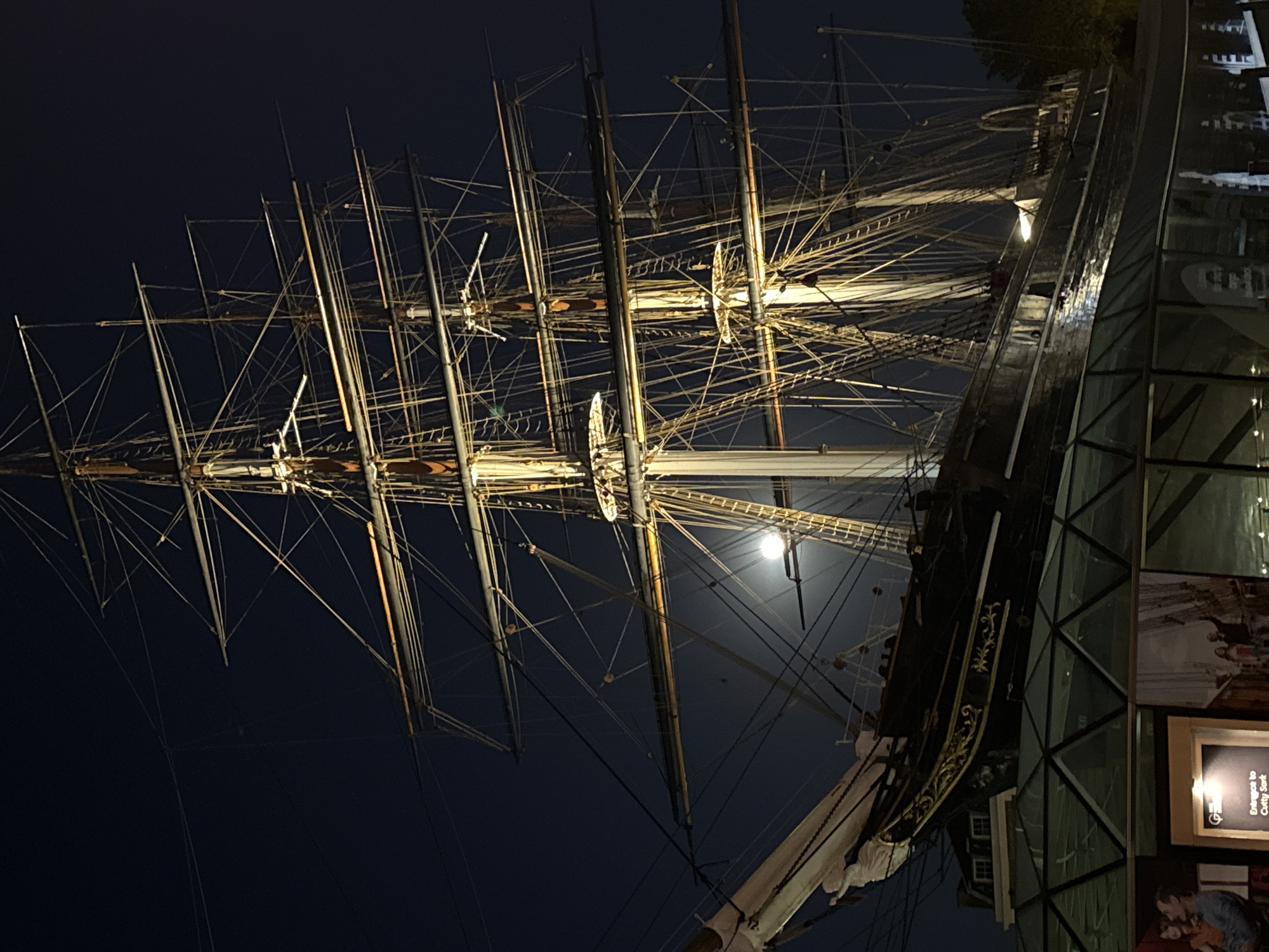 This striking nocturnal image captures the historic clipper ship, Cutty Sark, dramatically illuminated against a dark sky. The ship, with its numerous towering masts and intricate network of rigging, dominates the left and central portions of the frame. The masts are slender and tall, with yards extending horizontally, suggesting where sails would have been set. Many lines and ropes crisscross between the masts, creating a complex web. The ship's dark hull, with decorative gold accents near its stern, is visible on the right, curving upwards towards the viewer.
Behind the ship's rigging, a bright, full moon is visible, casting a soft glow and adding a natural light source to the scene. The time of day is clearly night, indicated by the dark sky and the prominent moon. The ship itself is brightly lit by artificial lights, highlighting its structure and the details of its rigging.
To the right of the ship, a modern glass and metal structure, likely a museum or visitor center, stands. The ship appears to be housed or displayed within or alongside this building. Inside the glass structure, promotional posters or displays are visible. One prominent black sign with a white frame clearly displays the text "Entrance to Cutty Sark", confirming the identity and location of the ship, which is preserved in Greenwich, London. Below this, part of another display showing a person is visible.
The scene conveys a sense of historical grandeur and preservation, with the ancient vessel contrasted against a contemporary architectural setting under the serene night sky. No explicit human activity is depicted; the focus is on the ship as a static, preserved artifact. The weather appears clear, allowing for the unobstructed view of the moon.