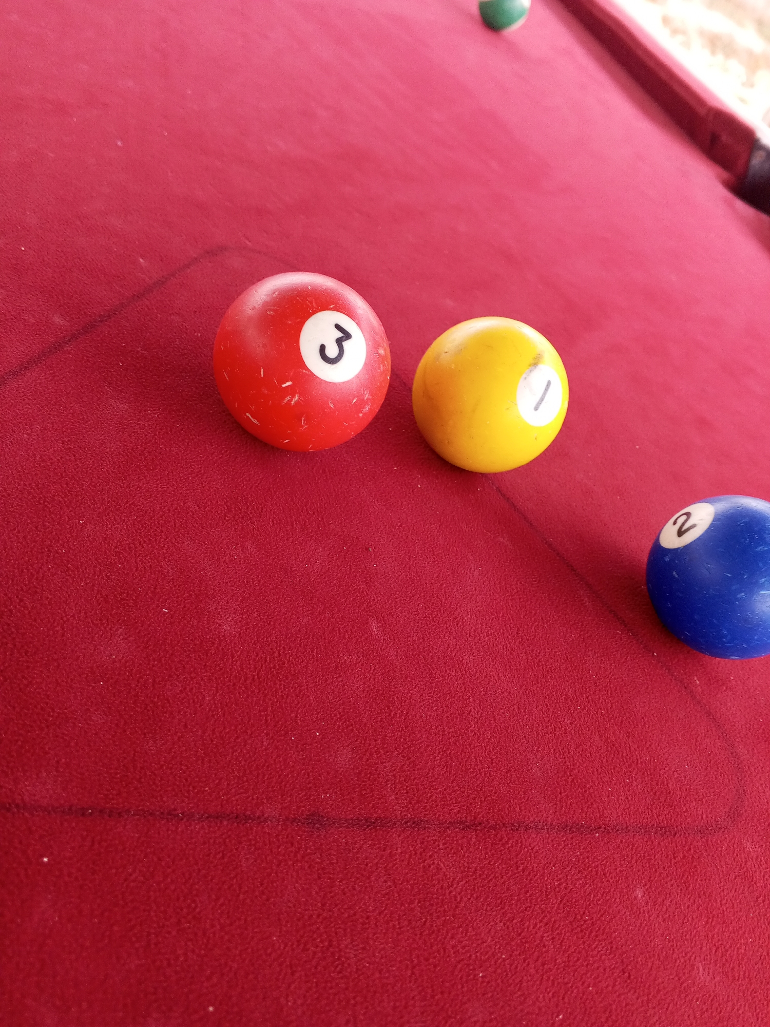 A close-up, high-angle shot shows a portion of a red felt billiard table. Three billiard balls are visible. On the left, a red ball with a white circle containing the number "3" is slightly out of focus. To its right, a yellow ball with a white circle containing the number "1" is also slightly out of focus. In the lower right corner, a blue ball with a white circle containing the number "2" is in focus. A faint black line, likely a chalk mark or part of the table's layout, runs horizontally across the image. A small portion of a green billiard ball is visible in the upper background, out of focus and near the edge of the table. The lighting suggests an outdoor or semi-outdoor setting, with natural light illuminating the scene. The table is positioned on an unseen surface, with what appears to be foliage or dry ground visible in the blurred background.