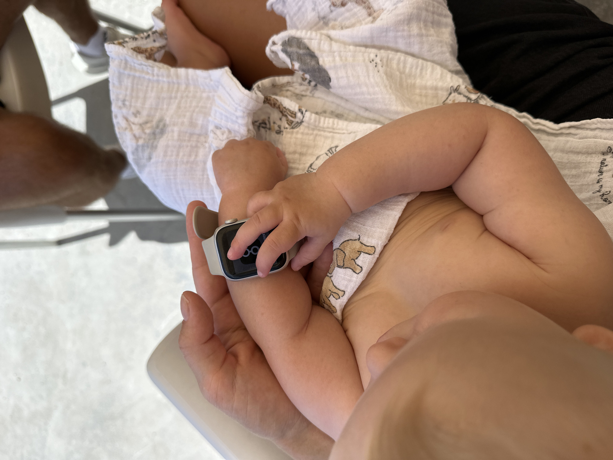 This media file captures a close-up view of a baby's arm and hand interacting with a smartwatch. The baby's small hand, with chubby fingers, is touching the screen of a silver Apple Watch. The watch face displays a digital time of "09:06" and a circular activity progress indicator.
The baby is wrapped in a white muslin cloth adorned with a subtle pattern of brown elephants. The baby's skin appears smooth and soft, with a hint of redness on the arm. Partially visible in the background are adult legs, one wearing white sneakers, suggesting an outdoor setting. The ground appears to be a light-colored, textured surface, possibly concrete or pavement, with shadows cast by an unseen object, indicating daylight.
The overall impression is one of gentle interaction and a moment of discovery. The focus on the baby's hand and the smartwatch creates a sense of curiosity and highlights the juxtaposition of technology with the innocence of infancy. The natural light and relaxed pose suggest a casual, perhaps familial, setting. While the exact location is not explicitly shown, the provided context indicates Budva, Montenegro. The image evokes a peaceful and heartwarming scene.
