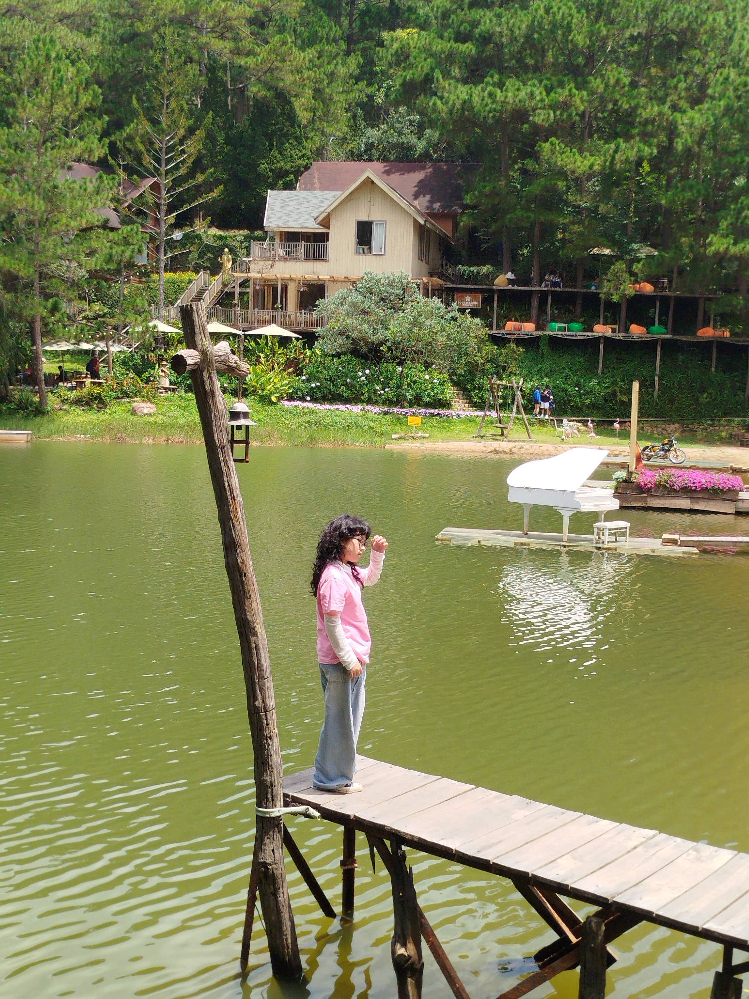 The image depicts a girl standing on a wooden pier extending into a calm, green body of water, likely Cam Ly Lake in Da Lat, Vietnam. Behind her, a dense forest of pine trees lines the shore, providing a backdrop to several elevated wooden structures housing buildings and seating areas with beanbag chairs. In the water, a white grand piano rests on a floating platform, accompanied by a piano bench. The pier itself is rustic, with visible wooden supports and planks. The girl is wearing a pink long-sleeved shirt and light blue jeans. The overall scene suggests a serene and scenic tourist destination.
