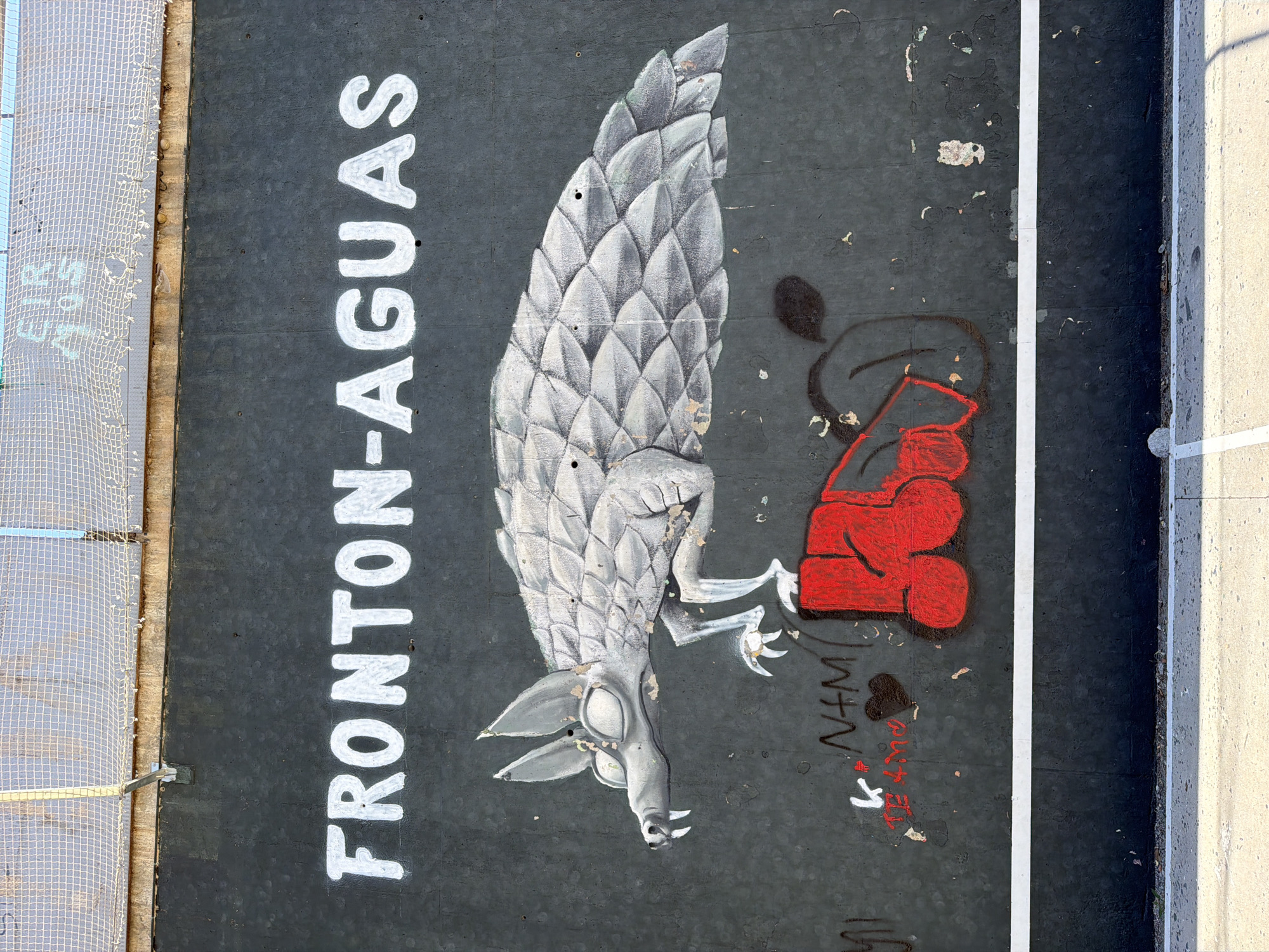 The image shows a mural painted on a dark grey wall. The mural depicts a stylized pangolin-like creature with scales, sharp teeth, and large ears. It is positioned above red graffiti that resembles a stylized hand or fist. White text reading "FRONTON-AGUAS" is painted prominently on the upper part of the wall. The wall appears weathered and has some peeling paint and other graffiti marks. A white horizontal line runs across the lower section of the wall, separating it from a concrete ground surface. A chain-link fence is visible at the very top of the image. The overall scene suggests an outdoor public space, likely a wall in the city of l'Hospitalet de Llobregat, Spain.