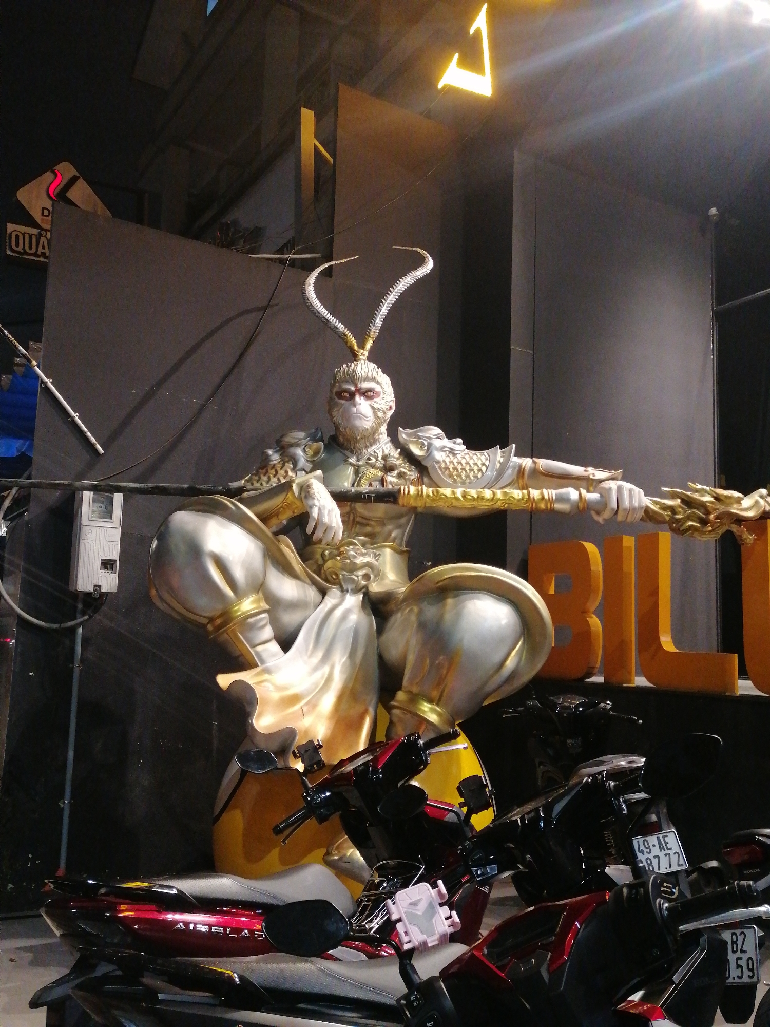 This nighttime image from Da Lat, Vietnam, features a large, elaborately detailed statue of what appears to be the Monkey King (Sun Wukong) at its center. The statue is depicted in a powerful, seated stance, holding a long, golden staff. It is adorned with intricate golden and silver armor, and its face is monkey-like with white fur, prominent red markings around the eyes, and large, stylized horns or antlers atop its head.
The setting is an outdoor or semi-outdoor commercial area, characterized by dark walls and structures in the background, illuminated by artificial light sources. To the left of the statue, a white electrical meter box is mounted on the wall, with a white pole leaning against it. A sign on the left wall shows the partially visible text "QUA" and a red, wavy arrow design.
Several motorbikes and scooters are parked around the base of the statue. A red scooter on the left clearly displays the brand name "AIRBLADE". Two visible license plates on other scooters read "49-AE 877.72" and "82 .59". To the right of the statue, large, golden block letters on the ground spell out "BI L", likely part of a larger word. Above, a bright yellow neon-like sign, shaped like a stylized "7" or "L", stands out against the dark sky. The scene is static, capturing the imposing statue and the surrounding elements under artificial illumination.