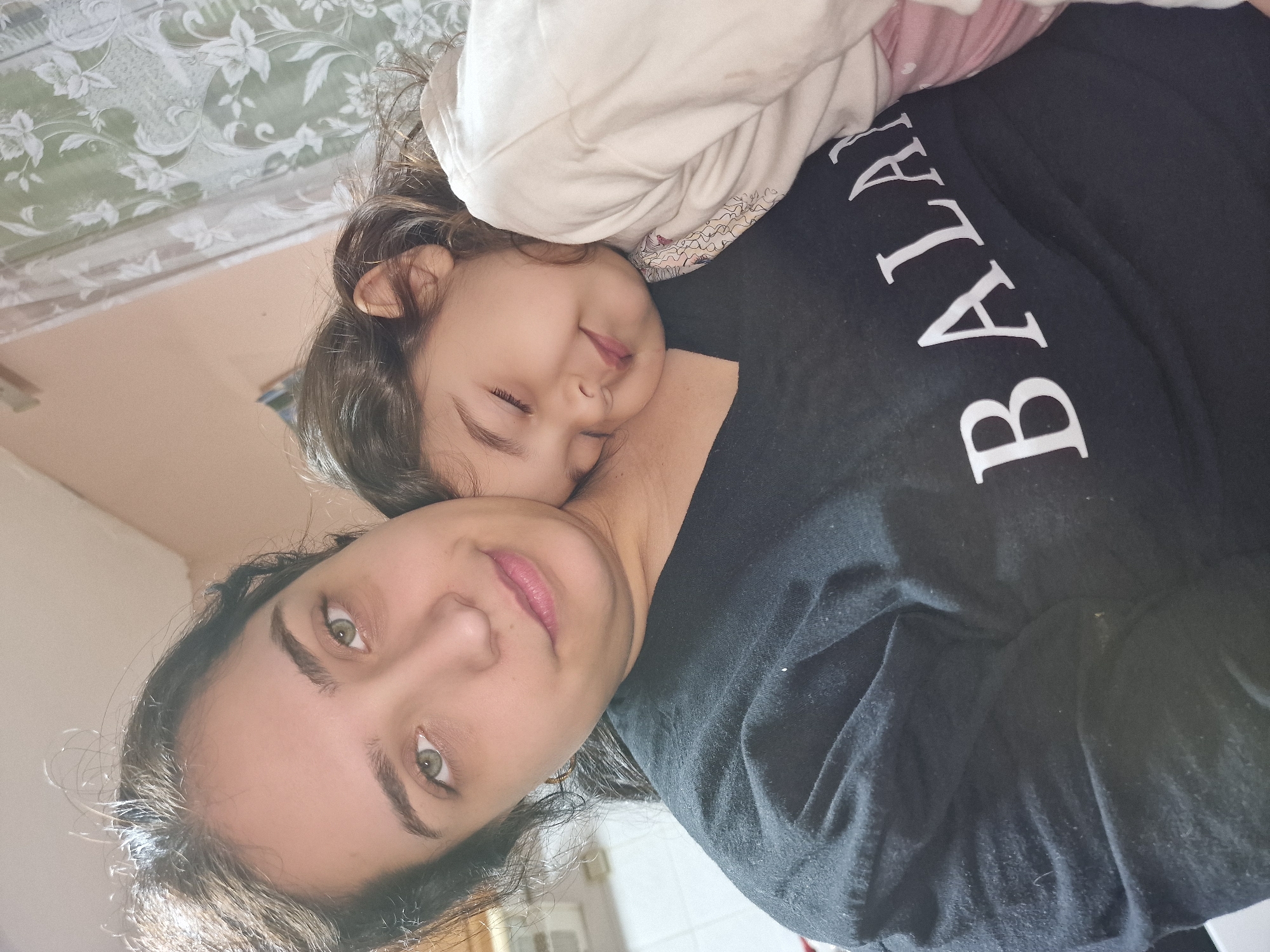 A medium shot shows a woman holding a sleeping child. The woman is fair-skinned with dark curly hair and green eyes. She is wearing a black t-shirt with white lettering. The child, also fair-skinned with dark curly hair, is nestled against the woman's chest and has their eyes closed. They are resting on a white pillow. The background is blurry, but a floral pattern on the bedspread is visible, as is the corner of a white wall. The location is in the city of Unknown, Unknown.