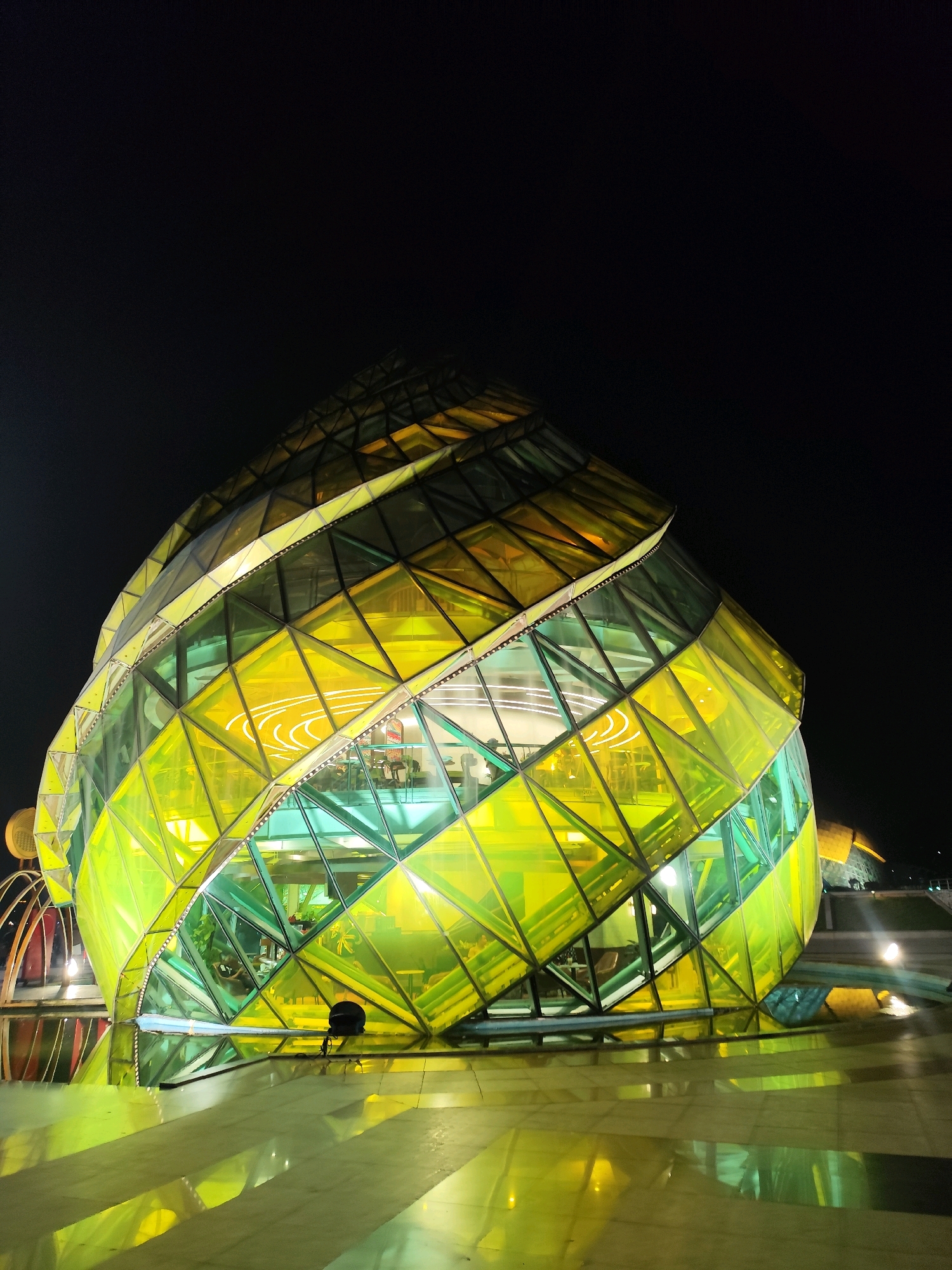 A brightly lit, multi-faceted glass building, resembling a large, segmented orb with a spiral design, stands against a dark night sky. The glass panels are a vibrant yellow and green, reflecting light and casting illuminated patterns on the wet, paved ground in front of it. Inside the translucent structure, there are visible lights and the silhouettes of people and furniture, suggesting it may be a restaurant or cafe. The building is situated in an open, outdoor area, possibly a plaza or promenade, with the reflection of the building and lights visible on the polished surface of the ground. In the background, a few distant lights and a dark, sloping structure can be observed. This is the iconic Da Lat Flower Garden building, also known as the Da Lat Pedagogical College or "Big C" building, located in Da Lat, Vietnam.