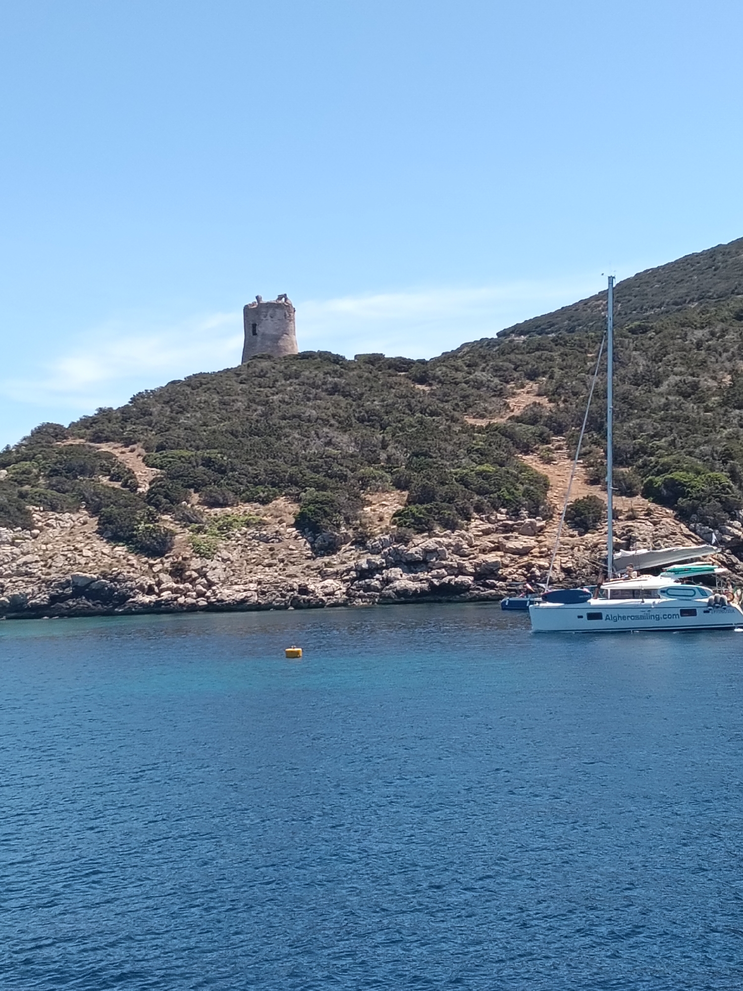 The image depicts a sunny day in Alghero, Italy, with a clear blue sky and calm, deep blue water. In the foreground, the sea is calm, with a yellow buoy visible in the mid-distance. A white catamaran with two hulls is moored in the water, displaying the text "Algherosailing.com" on its side. The catamaran has several people on board. Behind the boat and the water, a steep, rocky hill covered in dense, dark green vegetation rises up. At the summit of the hill stands an ancient, weathered stone tower, its crenellated top partially broken. The tower appears to be a historical landmark. The overall scene conveys a sense of leisure and a coastal environment.