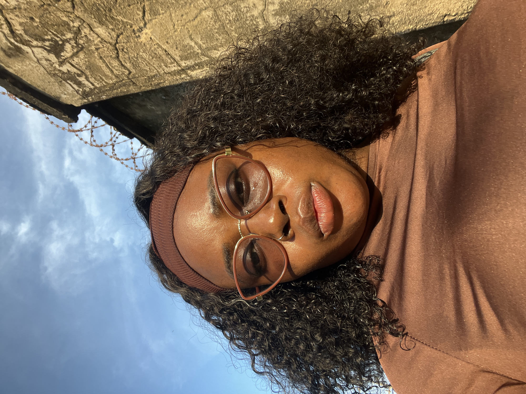 A young woman with dark curly hair and light brown skin is shown in a close-up, low-angle shot. She is wearing a brown headband and large, light brown-tinted glasses with a thin gold frame. A subtle nose ring is visible. Her lips are parted, and her expression is neutral. She is wearing a shiny, brown, long-sleeved top.
The background consists of a textured, beige-colored concrete wall on the right, topped with coiled barbed wire. Above the wall and the woman's head, a blue sky with scattered clouds is visible. The lighting suggests it is daytime, with sunlight casting shadows across the woman's face and clothing.
The presence of the barbed wire atop the wall hints at a potentially secure or restricted area, common in various urban settings. While the image does not directly depict identifiable landmarks of Abuja, Nigeria, the style of the wall and the presence of barbed wire are consistent with security features found in residential, commercial, or governmental compounds within the city. The sky and general urban feel are also common to Abuja.