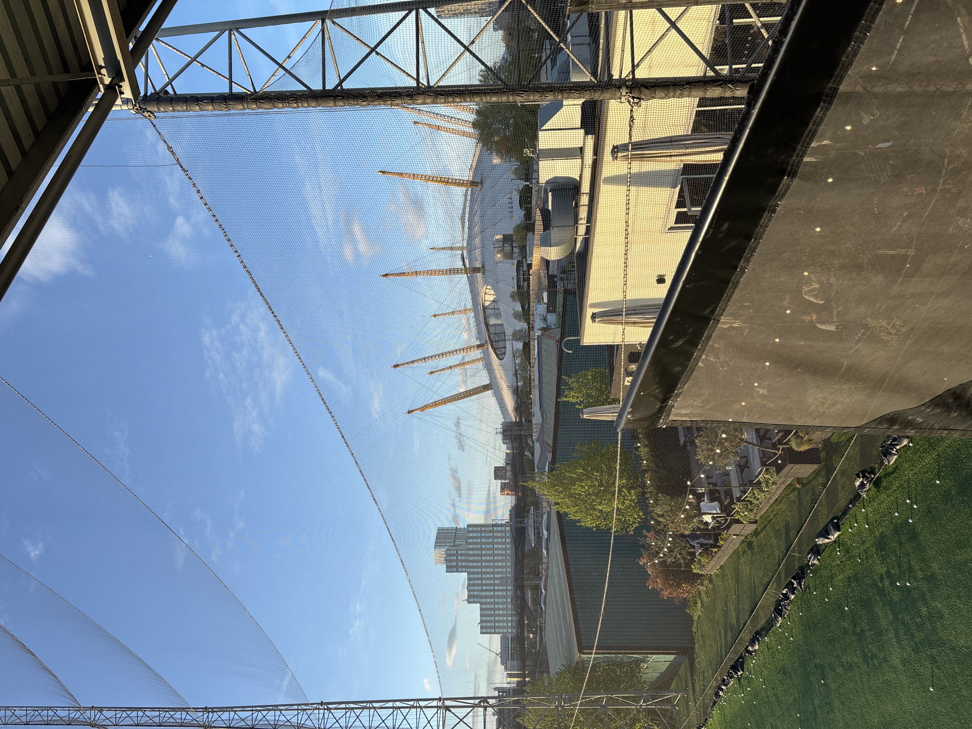 The original image is rotated 90 degrees clockwise. When viewed correctly, it presents an expansive outdoor scene from a golf driving range, offering a panoramic view of an urban landscape in London.
In the immediate foreground, metal structural beams and a dense, fine-mesh protective netting span across the upper and left portions of the frame, indicating an enclosure or safety barrier. Below this netting, towards the right side of the image, lies a vibrant green artificial turf area. This area is clearly a golf driving range, populated with numerous white golf balls placed on tees and several black golf bags neatly lined up along its edge. Adjacent to the driving range, further to the right, an inviting outdoor patio area is visible, featuring string lights, tables, and chairs, nestled amongst trees and bushes, suggesting a leisure or dining space.
The background is dominated by a clear, bright blue sky adorned with scattered white clouds, indicating pleasant weather. A prominent landmark in the middle distance, towards the left, is the iconic white domed structure of The O2 Arena, instantly recognizable by its distinctive yellow support masts. Beyond The O2 and stretching across the horizon, a cluster of modern high-rise buildings contributes to the urban skyline.
The lighting in the image suggests late afternoon or early evening, with the sun casting a warm glow across the scene. While no individuals are visible actively golfing or socializing, the setup of the driving range and patio clearly indicates a recreational and entertainment venue. No visible text is present in the image. The scene captures a dynamic blend of sports, leisure, and notable London architecture.
