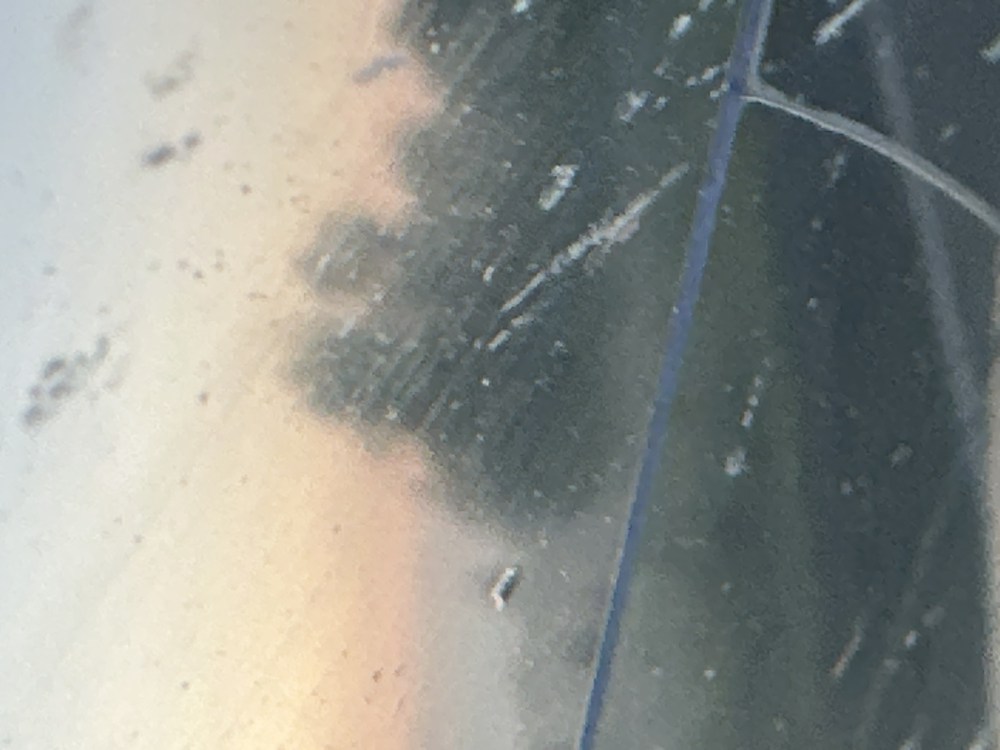 The image depicts a blurry, abstract scene with a gradient of colors suggesting a sunrise or sunset. The foreground appears to be a dirty or smudged surface, possibly a window, with streaks and water droplets. In the mid-ground, indistinct shapes of trees are visible against a soft, warm-toned sky. A thin, dark horizontal line runs across the mid-section of the image. The overall impression is one of an outdoor view through a soiled barrier, potentially captured at dawn or dusk. No specific city features or recognizable landmarks from Krapivinskiy, Russia are discernible due to the image's lack of clarity.