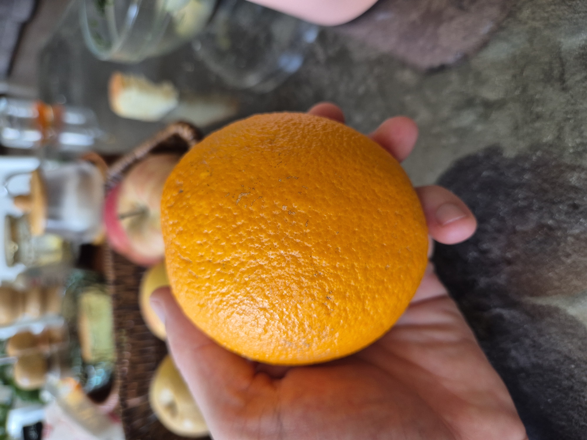 A close-up shot focuses on a hand holding a bright orange orange. The orange has a textured peel with small blemishes. In the blurred background, a dark-colored woven basket holds several fruits, including at least one apple. Other out-of-focus items like jars and a slice of cake are also visible on a reflective surface, possibly a table. The overall setting appears to be a domestic environment. There is no information about the location in Taldykorgan, Kazakhstan.