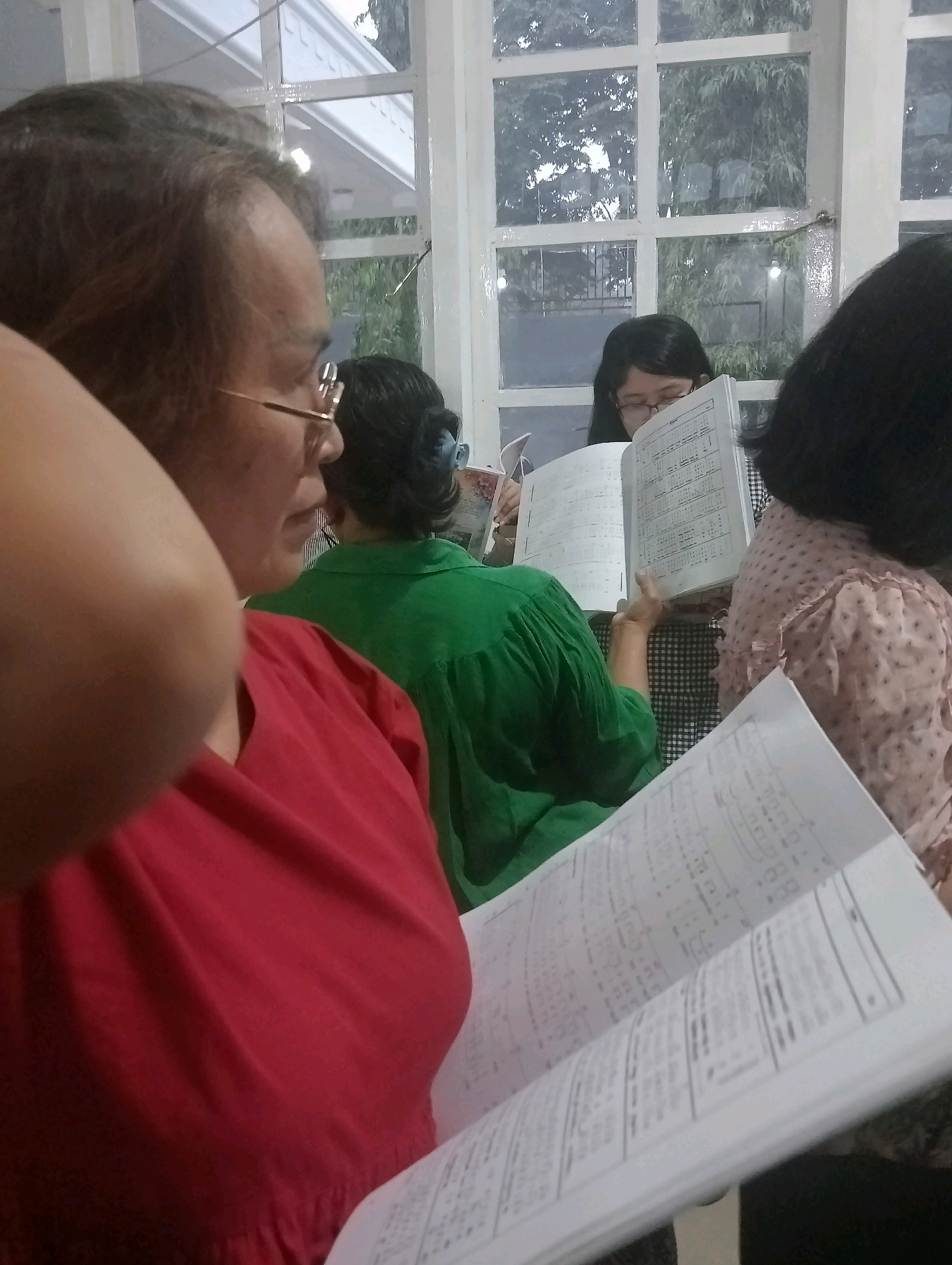 In Jambi, Indonesia, four women are seen gathered indoors, likely in a community hall or home, for a choir practice or similar musical activity. One woman, in the foreground and to the left, is wearing a red top and is in profile, her arm visible. Behind her, a woman in a green shirt faces away from the camera, holding an open book. To her right, another woman, wearing glasses, is looking down at an open book. The fourth woman, to the far right, is wearing a pink patterned top and is also looking at a book. The environment suggests a well-lit room with a large window revealing trees and foliage outside, possibly indicating a suburban or natural setting within Jambi. The women are engaged in reading from their respective books, suggesting a shared activity involving music or study.