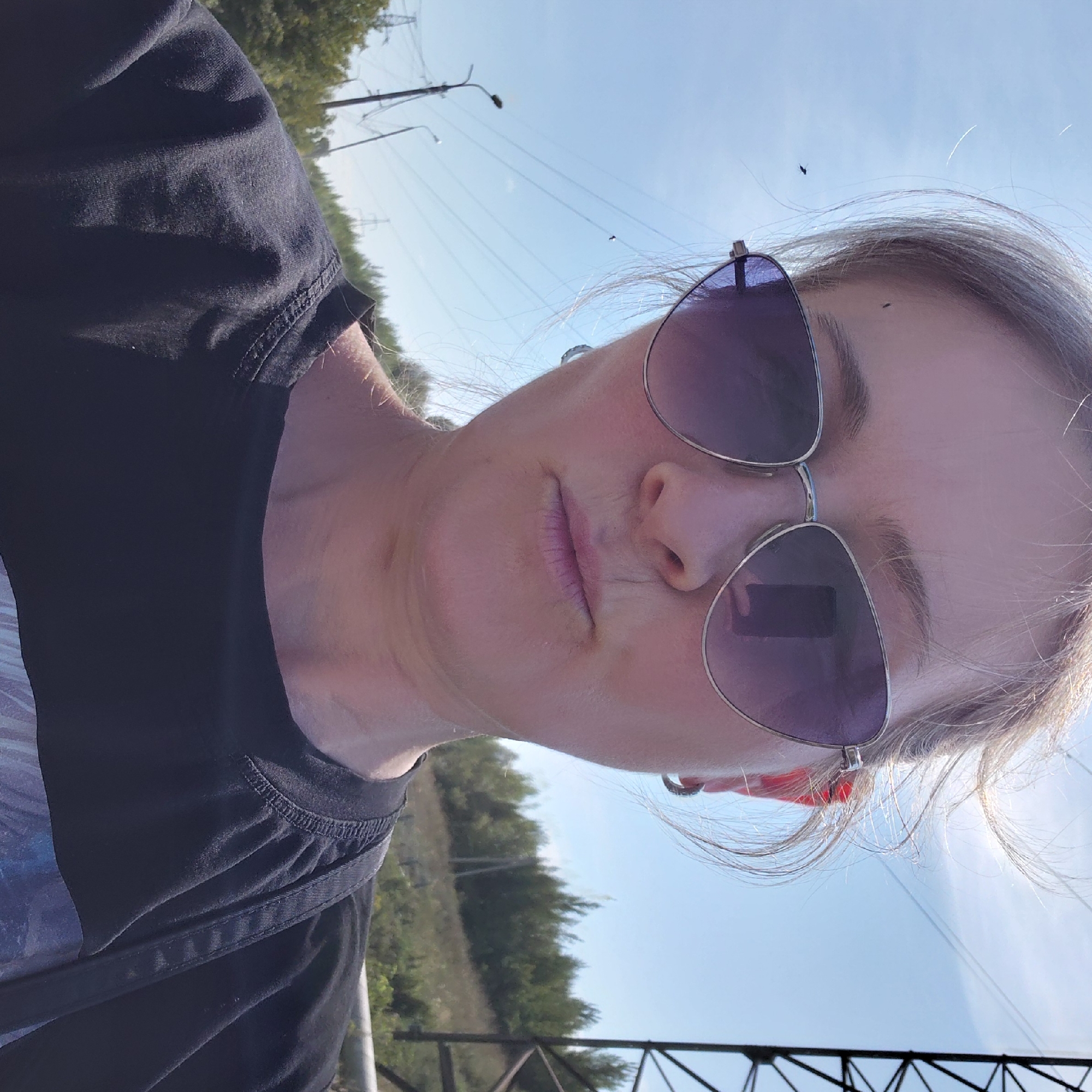 A woman wearing sunglasses and a black t-shirt takes a selfie outdoors. She is positioned in the center of the frame, with her face and upper body visible. Her hair is light-colored and blowing in the wind. The background shows a clear blue sky, trees, power lines, and a large industrial structure on the right. There are no other people or animals in the immediate vicinity. The lighting suggests it is daytime with bright sunlight. The location is not specified in the image or metadata.