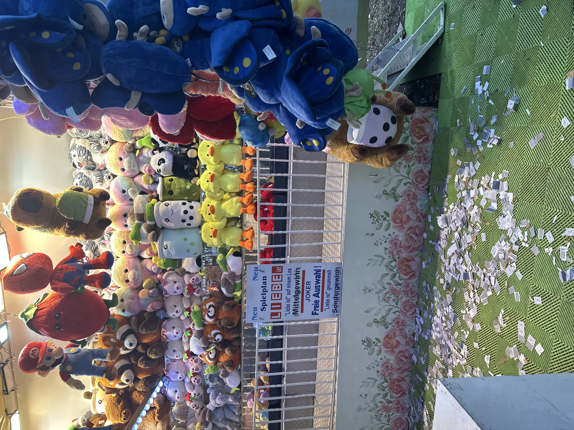 This image depicts a carnival game booth, likely in Bruchhausen-Vilsen, Germany, filled with plush toys. The booth features a tiered display of stuffed animals, including characters like Mario and Spiderman, as well as various animal figures and cartoon-themed plushies. In the foreground, a sign is prominently displayed with German text, indicating game rules and prizes, with the word "LIEBE" (love) central to the messaging. The ground in front of the booth is covered in numerous small, white slips of paper, suggesting discarded game tickets or coupons. A few larger plush toys hang from the top of the booth, and a brown teddy bear with a boba tea cup design on its belly hangs upside down from a wire. The background shows a floral wallpaper and a metal barrier.