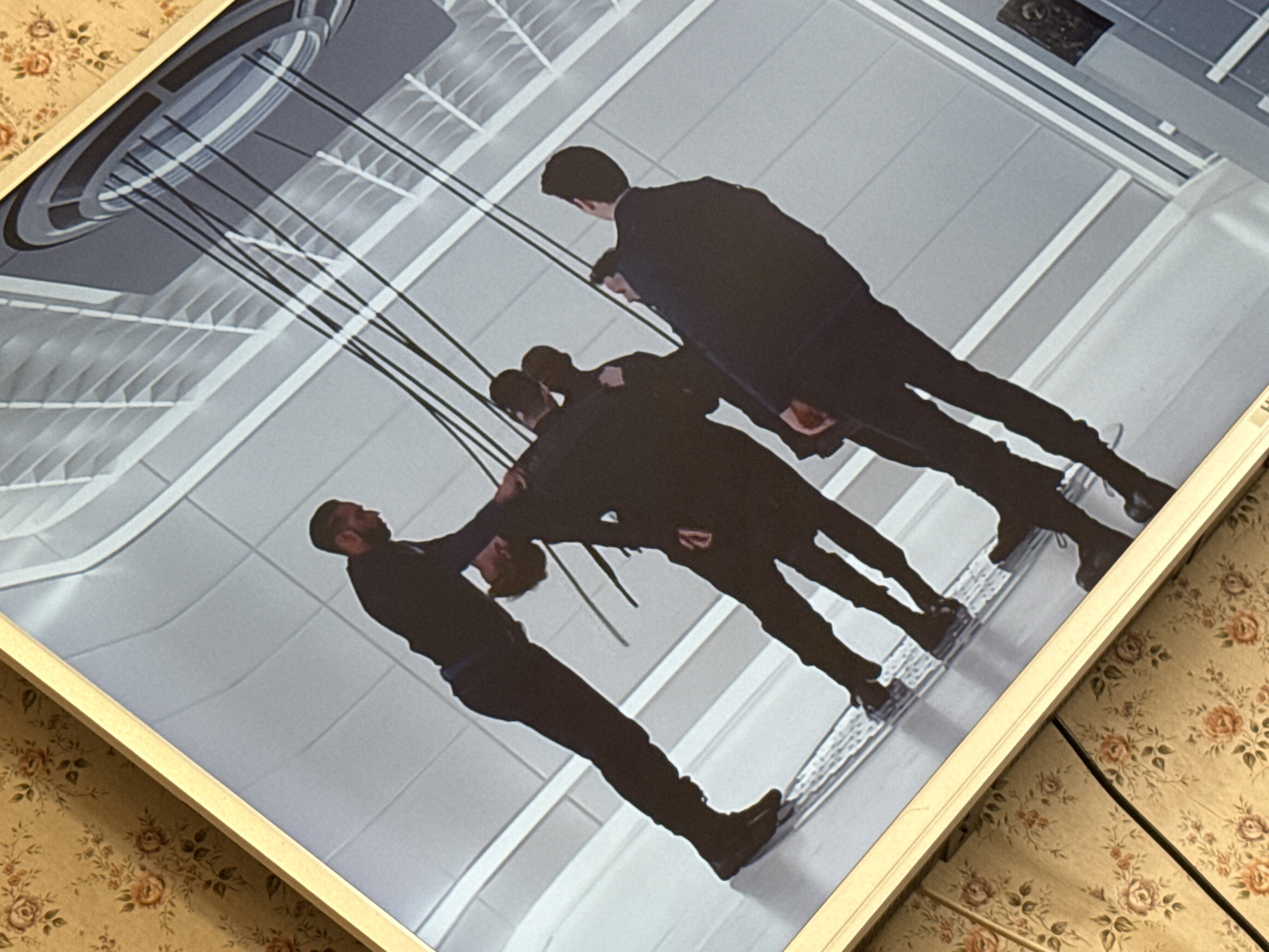 The image shows a framed photograph. The photograph depicts a scene indoors, featuring five men in dark suits standing on a shiny floor with a mirrored or reflective surface. The background consists of a grid-like ceiling pattern and a circular structure with hanging cables or wires. The photograph is framed with a white or light-colored frame and appears to be sitting on a surface covered with a floral patterned cloth.
The image does not contain sufficient information to identify the exact location in Mytishchi, Russia.
