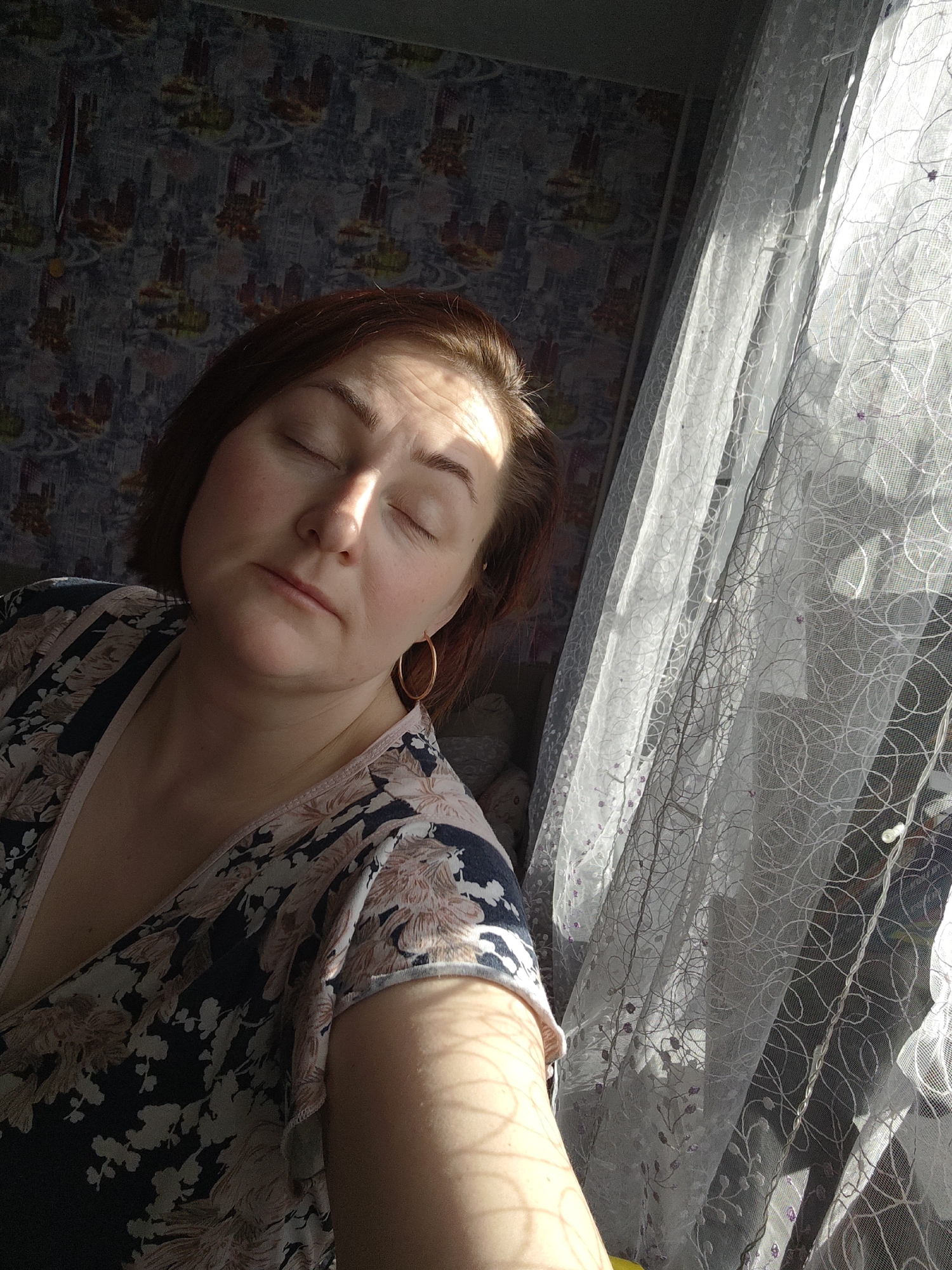 A medium shot captures a woman with reddish-brown hair and closed eyes, illuminated by sunlight. She wears a floral-patterned top. The background reveals a wallpapered wall with a dark, patterned design on the left, and a white, semi-transparent curtain, with sunlight streaming through, on the right. The scene is likely indoors, near a window. Markovskii, Russia is mentioned but no element in the image clearly connects it to this location.