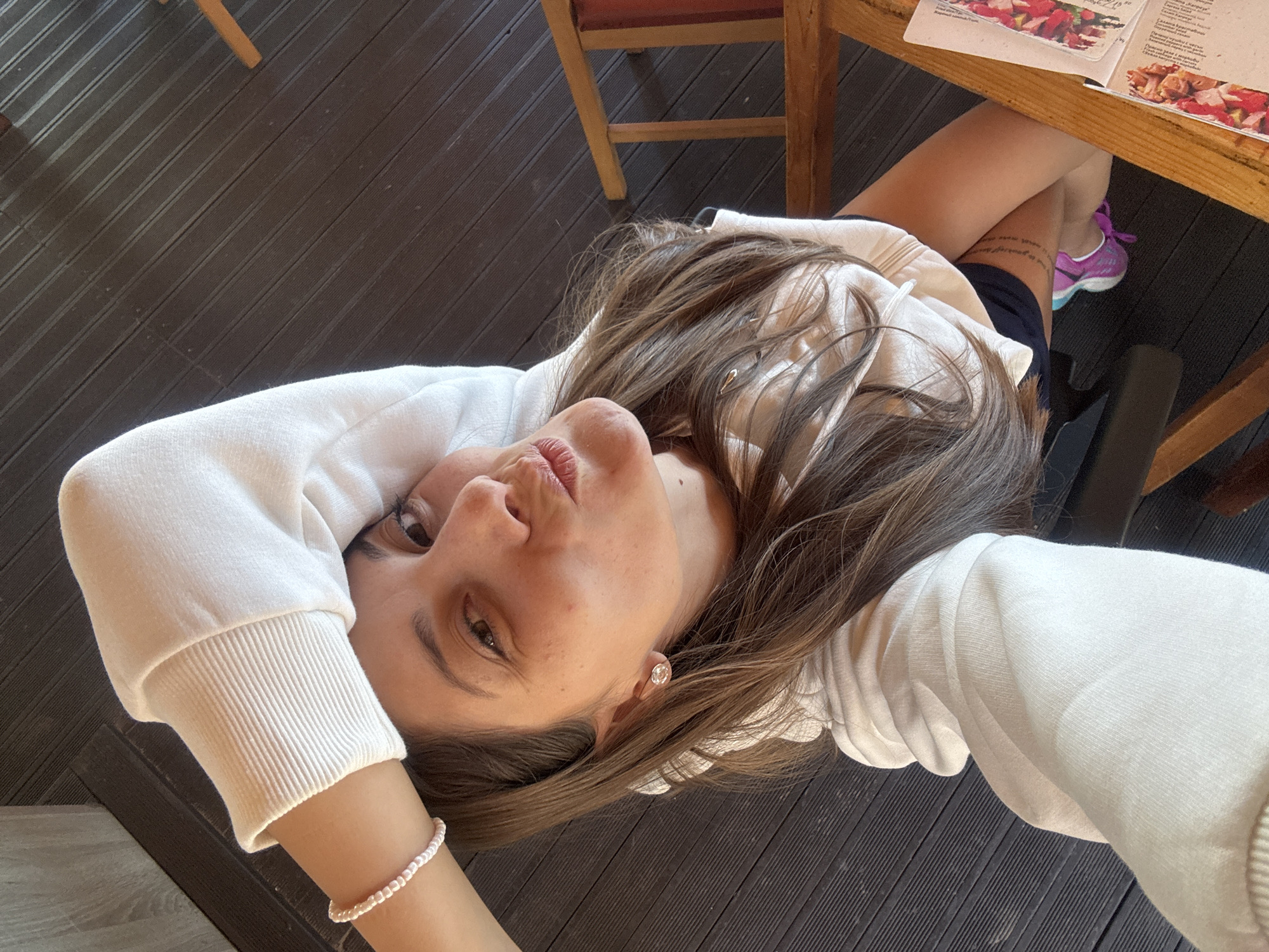 A woman with brown hair is posing for a selfie while seated at a wooden table. She is wearing a white sweatshirt and a pearl bracelet on her left wrist. Her right leg, clad in dark shorts and pink athletic shoes, extends under the table, where a menu is visible. The location appears to be in Sozopol, Bulgaria, based on the menu. The flooring is a dark wood-like material, possibly a deck.