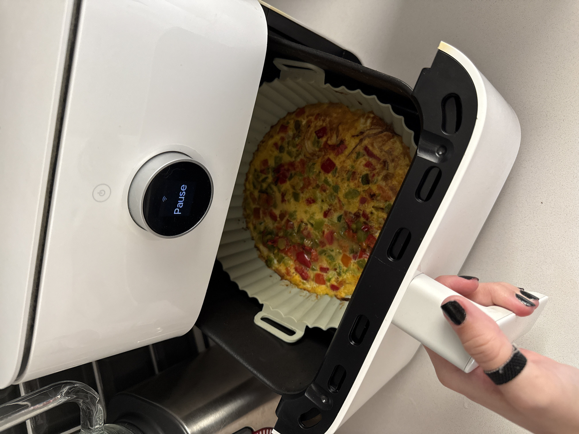 The image shows an opened white air fryer with a round, baked omelet in a silicone mold inside the drawer. The air fryer is likely in a kitchen, suggested by the countertop background. The air fryer's control panel is visible, displaying the word "Pause." A hand with black nail polish and a dark ring is holding the air fryer drawer handle. The location (Barcelona, Spain) cannot be determined from the provided image.