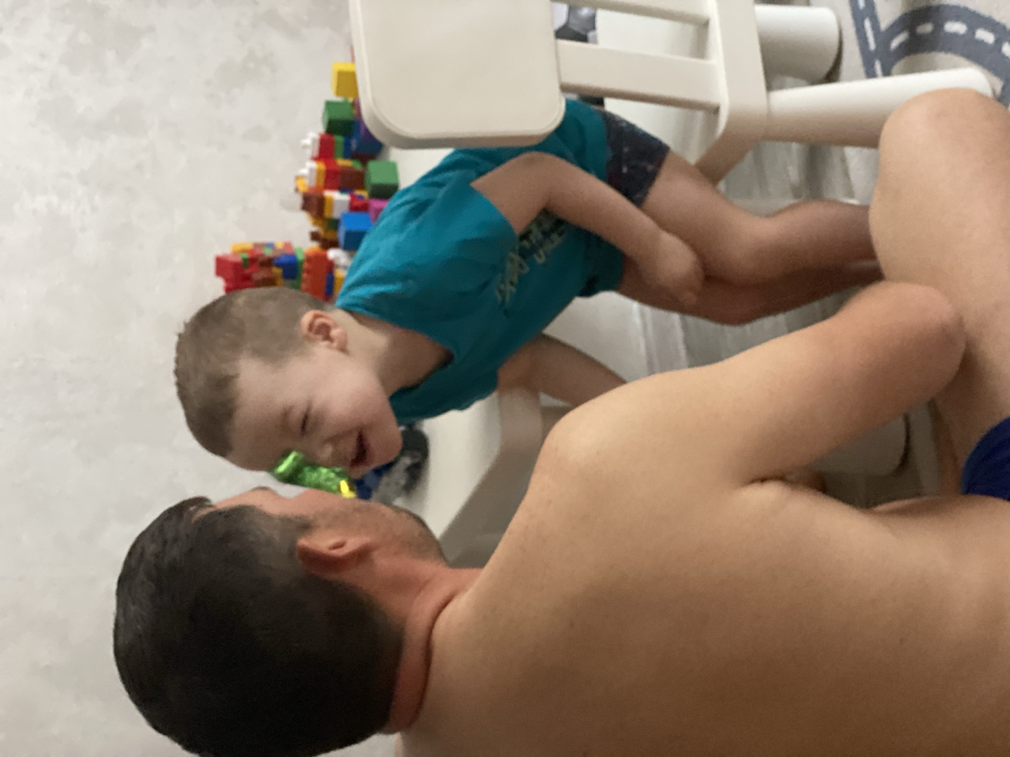 A man lies on his back while a child leans over him, likely interacting playfully. The man is shirtless and the child is wearing a blue shirt and dark shorts. The child appears to be laughing. A white chair with building blocks visible behind it is present. The location is not discernible from the image.