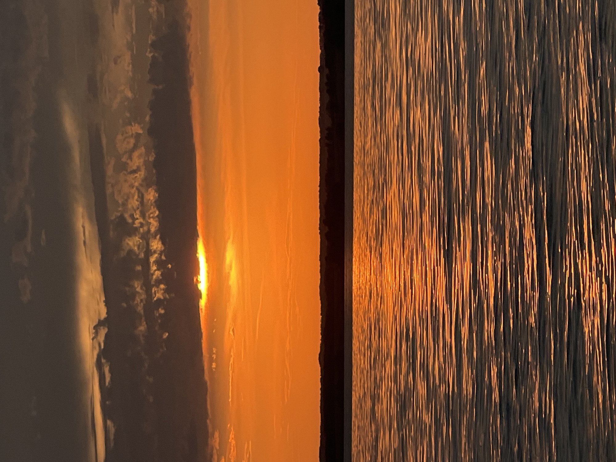 A sunset over a body of water is depicted. The sun is partially obscured by dark clouds, casting a bright orange glow across the sky. The water reflects the orange light, creating ripples of color. A dark, silhouetted treeline is visible in the distance. The scene is tranquil and appears to be a natural landscape, possibly near Beregovoe, Russia, given the context. There are no people or specific objects visible besides the natural elements.
