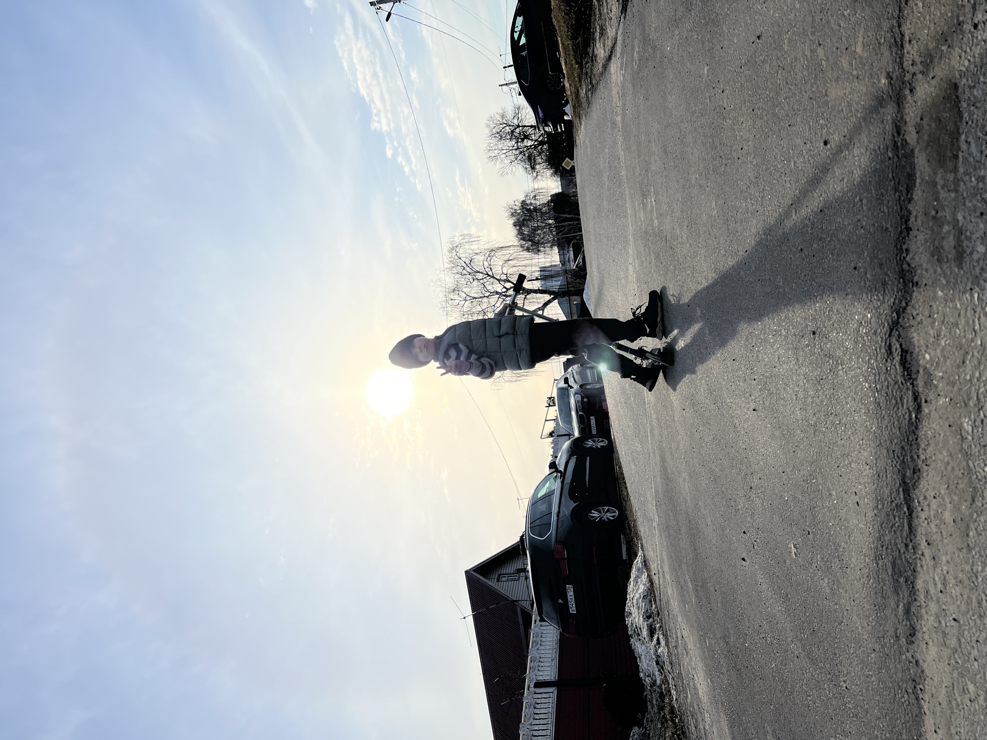 A person is standing on the edge of a road in Kazan, Russia, with cars parked along the side. The person is wearing a dark beanie, light-colored jacket, and dark pants. The sun is brightly shining behind the person, casting a shadow on the road. The road surface is asphalt. A snow-covered area is visible to the right of the road. Several cars are parked along the road, next to a red brick building with a pointed roof. Trees are in the background. Overhead power lines are visible in the sky.