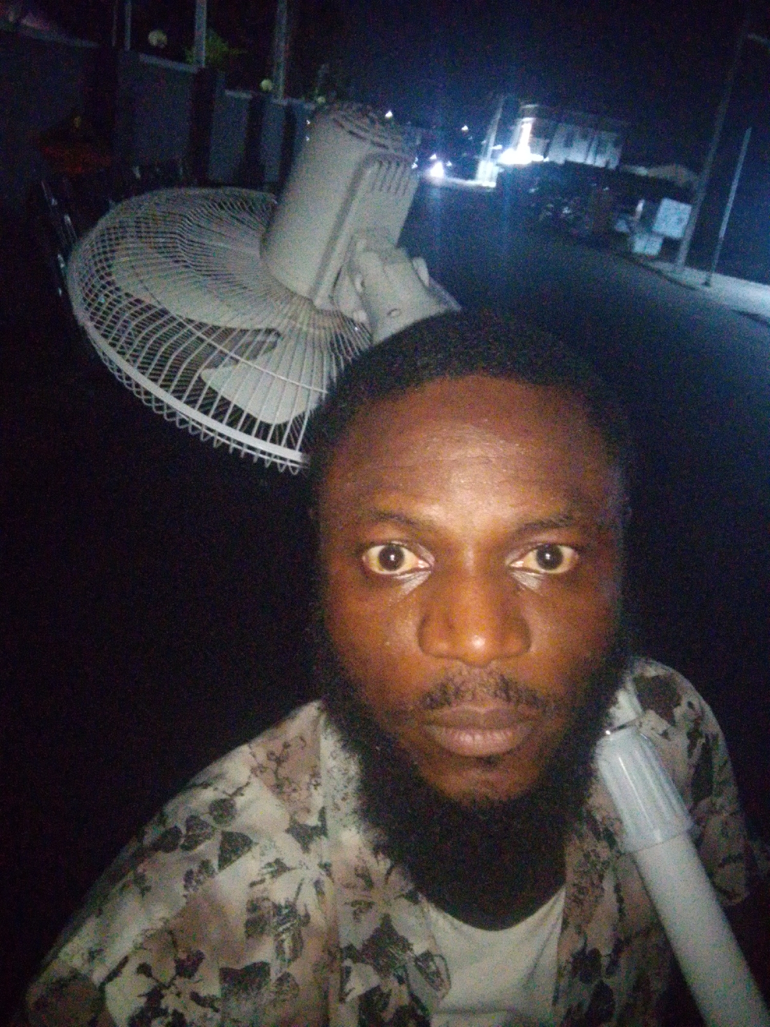 A person is holding a fan outdoors at night in a city. The person has a beard and is wearing a patterned shirt. The fan is white and tilted, and a portion of the fan's stand is visible. Streetlights and building lights are visible in the background.